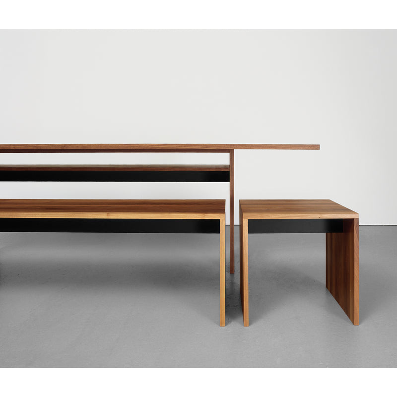 The Sanktjohanser Format Bench, part of a minimalist dining set made from solid wood, includes a long rectangular table and a smaller square table with sleek lines. The set is showcased on a smooth light gray floor against a plain white wall.