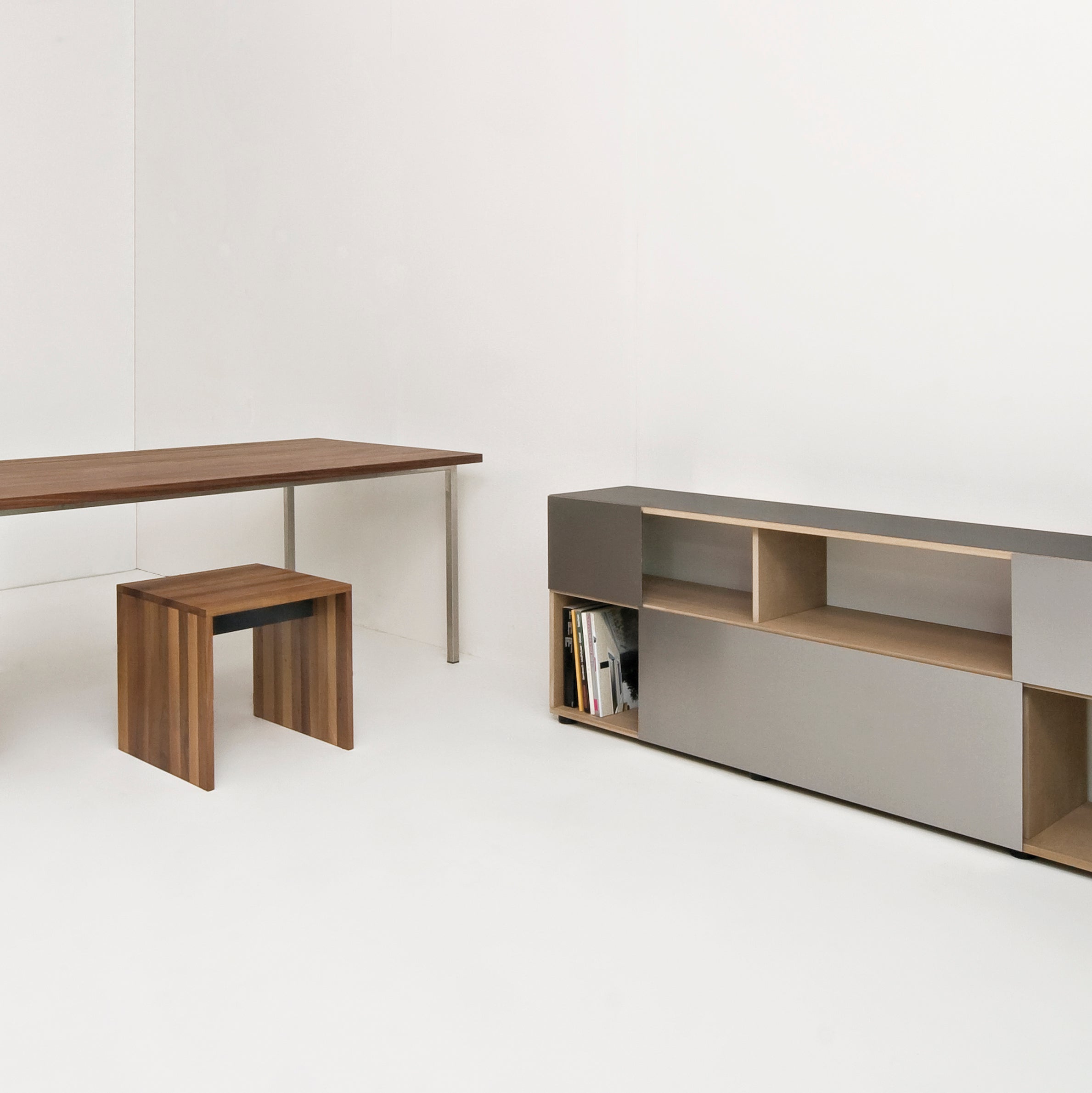A minimalist room with a wooden desk, matching stool, and modern cabinet showcasing open shelves with books maintains a neutral, uncluttered look. The Sanktjohanser Format Bench adds elegance with its impeccable solid wood finishes.