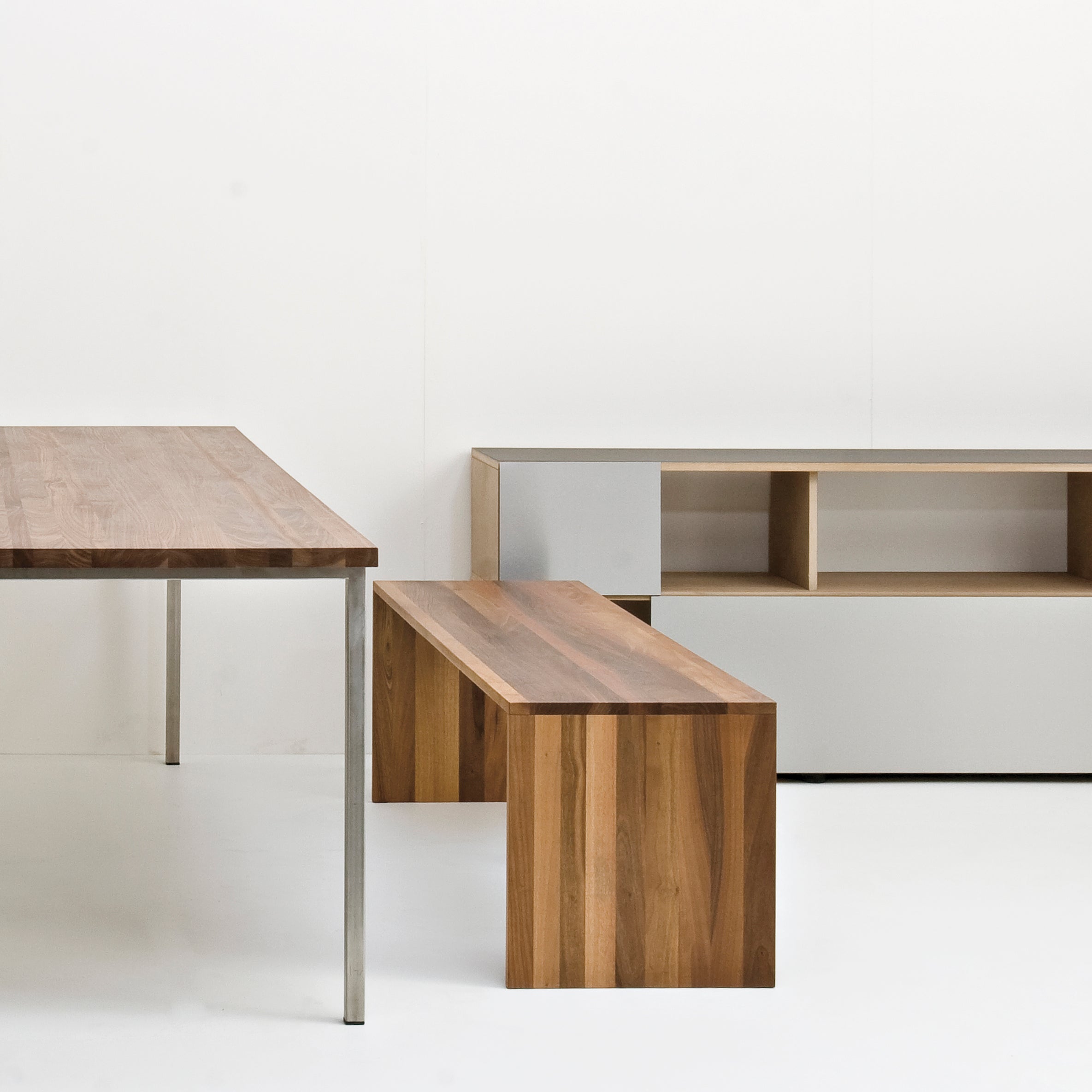 The Sanktjohanser Format Bench, with its clean lines, complements a minimalist setup featuring a wooden table with metallic legs and a cubical storage unit with open shelves against a plain white background.