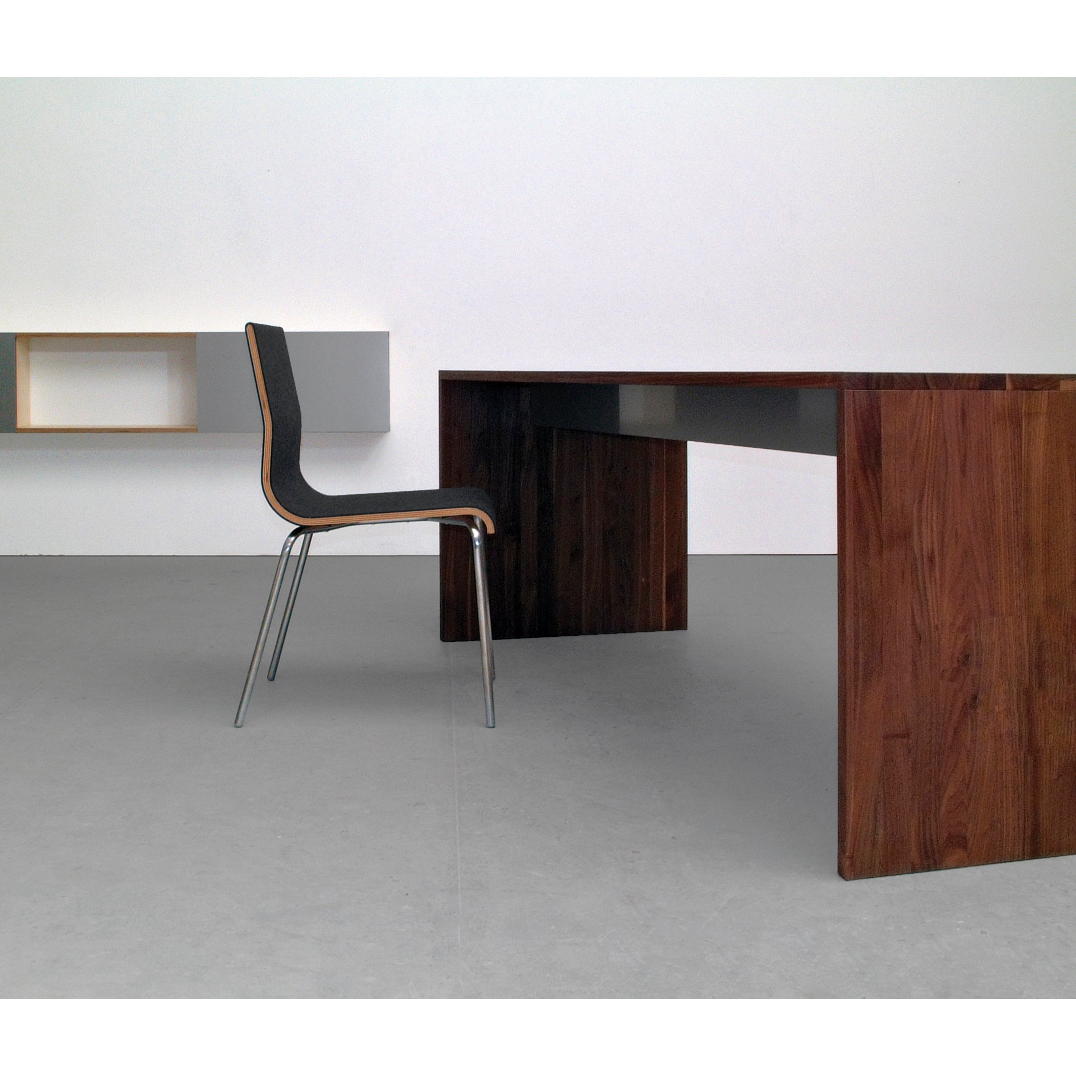 In a modern, minimalist office setting, the Sanktjohanser Format Table pairs with a sleek black cushioned chair on a gray floor. A simple wall-mounted shelf in the background enhances the clean aesthetic.