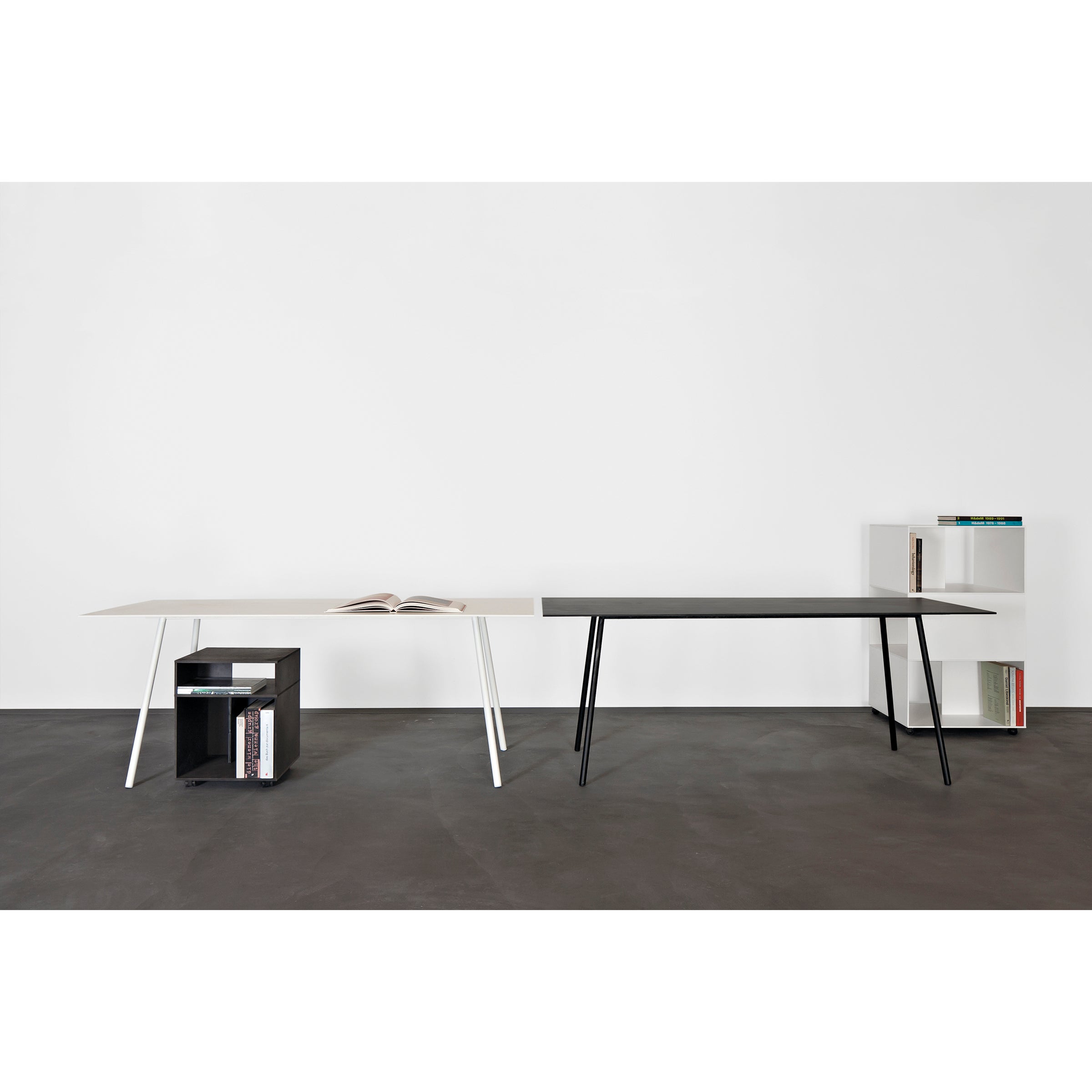 Two modern minimalist Kontext Tables by Sanktjohanser stand against a white wall. One is white with a black cube bookshelf underneath, made from durable round-bar steel. The other is black with a white bookshelf beside it. Both tables rest elegantly on the gray floor and offer custom sizes.