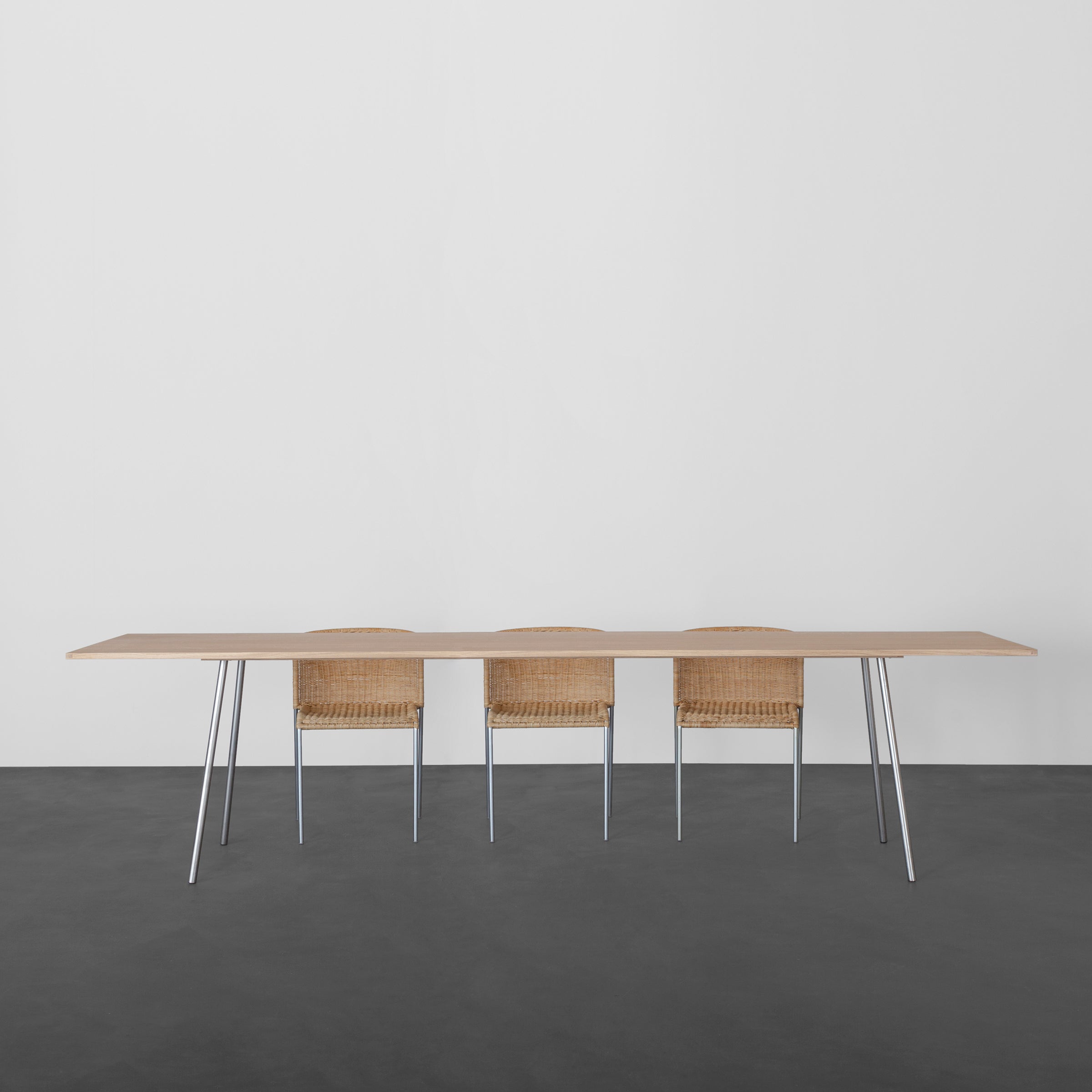 For a minimalist setup, consider the Kontext Table by Sanktjohanser with slender round-bar steel legs, paired with three woven chairs. The ensemble sits perfectly on a dark floor against a white wall. Custom sizes ensure an ideal fit for your space.
