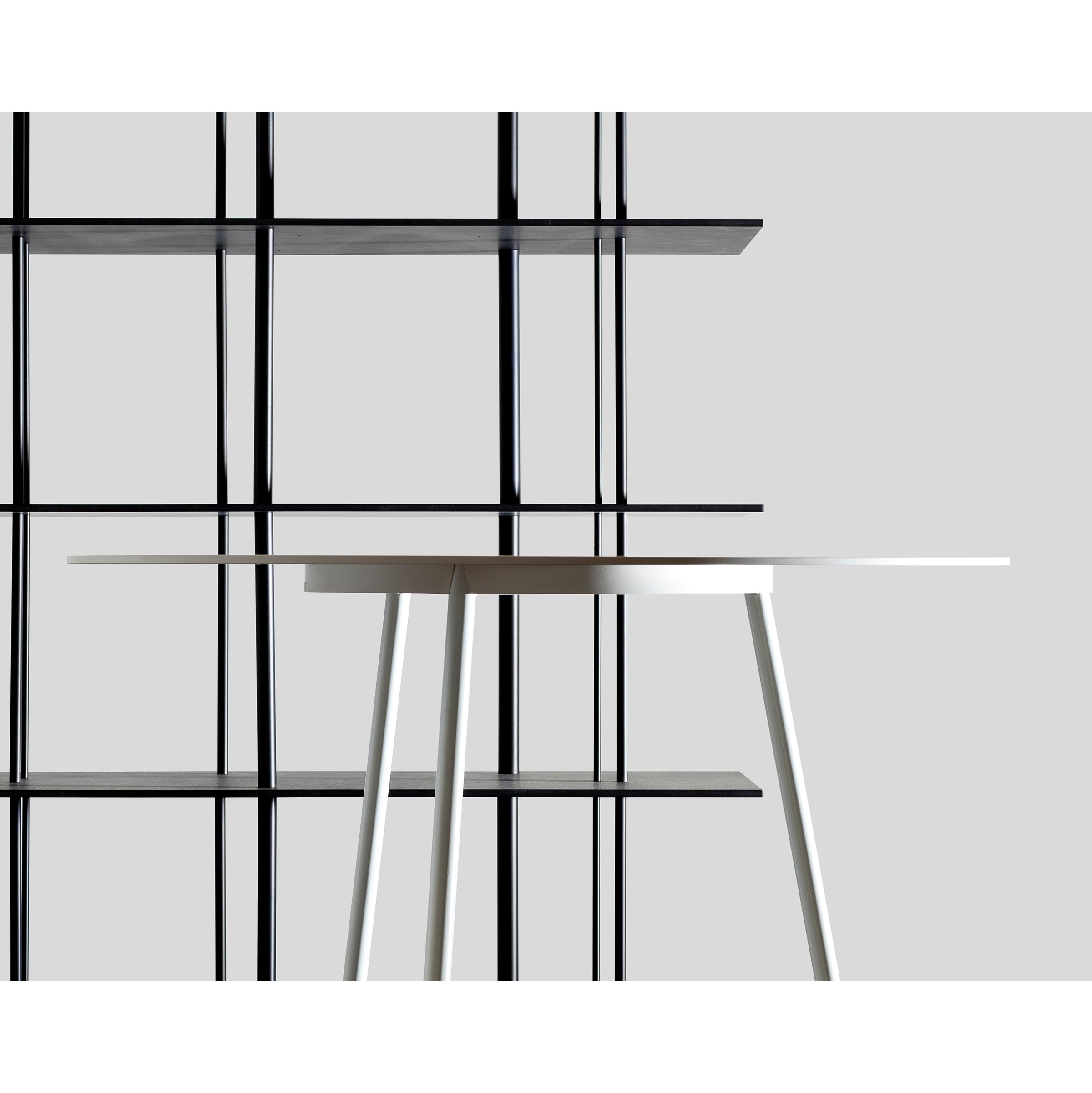 A minimalist scene features the Kontext Table by Sanktjohanser, crafted from solid wood, against a black geometric shelving unit made of round-bar steel. Set against a light gray background, its modern lines and custom sizes contrast strikingly in color and form.