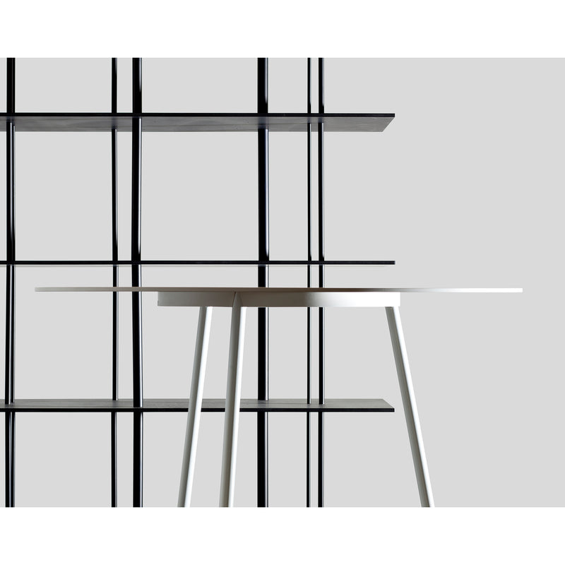 A minimalist scene features the Kontext Table by Sanktjohanser, crafted from solid wood, against a black geometric shelving unit made of round-bar steel. Set against a light gray background, its modern lines and custom sizes contrast strikingly in color and form.
