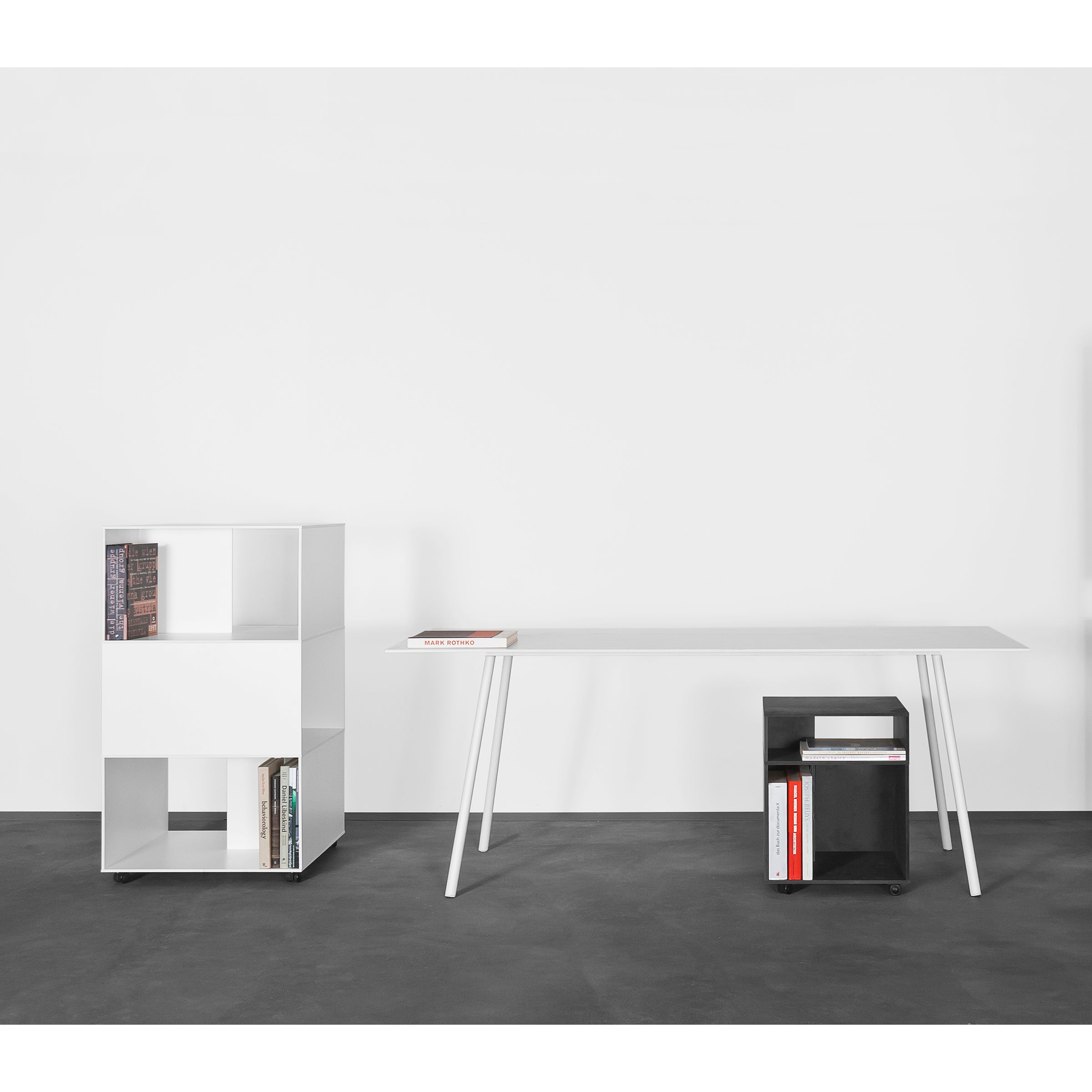 A minimalist office setup features a sleek Kontext Table by Sanktjohanser with a few books on top and a small, custom-sized black shelf below. A cube-shaped white bookshelf, partially filled with books, stands on the left against a plain white wall and dark floor.