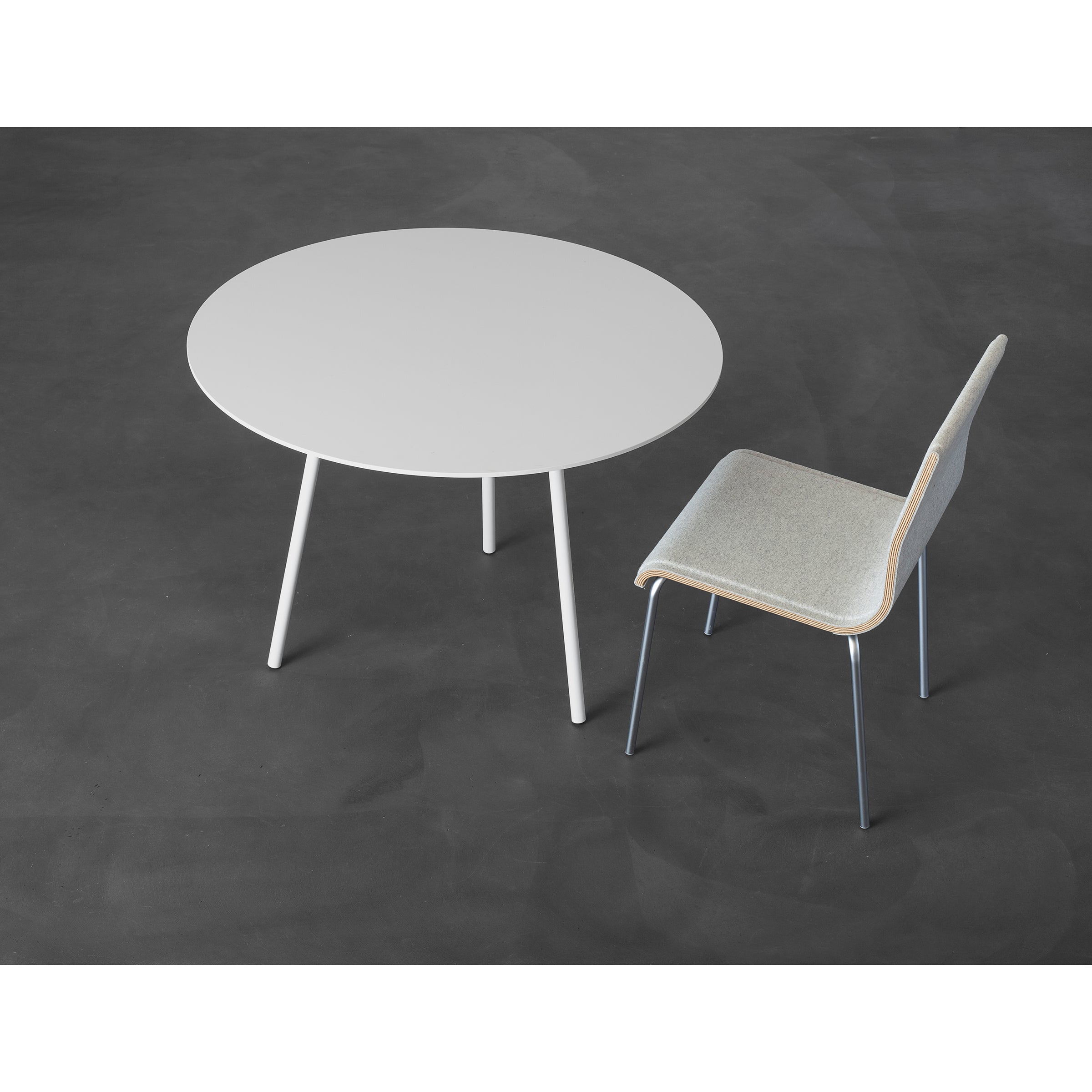 The Kontext Table by Sanktjohanser is a minimalist gray, round three-legged table made of round-bar steel. Its paired with a simple gray chair with a wooden backrest and metal legs. Custom sizes are offered to fit your space, all beautifully set on a dark gray floor.
