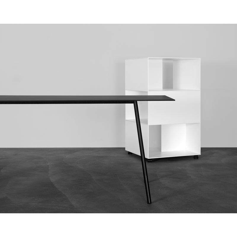 In a minimalist office, the Kontext Table from Sanktjohanser, featuring sleek black with thin, angled round-bar steel legs, is central. A white geometric shelving unit on wheels enhances functionality against the plain gray wall and dark floor. Custom sizes ensure a perfect fit for any space.