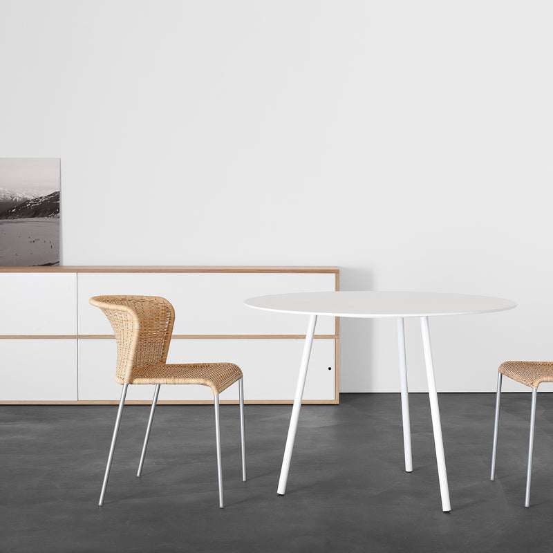 A minimalist room showcases a round, white Kontext Table by Sanktjohanser, two wicker chairs, and a wood-accented sideboard. A landscape painting subtly adorns the sideboard, while the dark floor elegantly contrasts with light walls and furniture.
