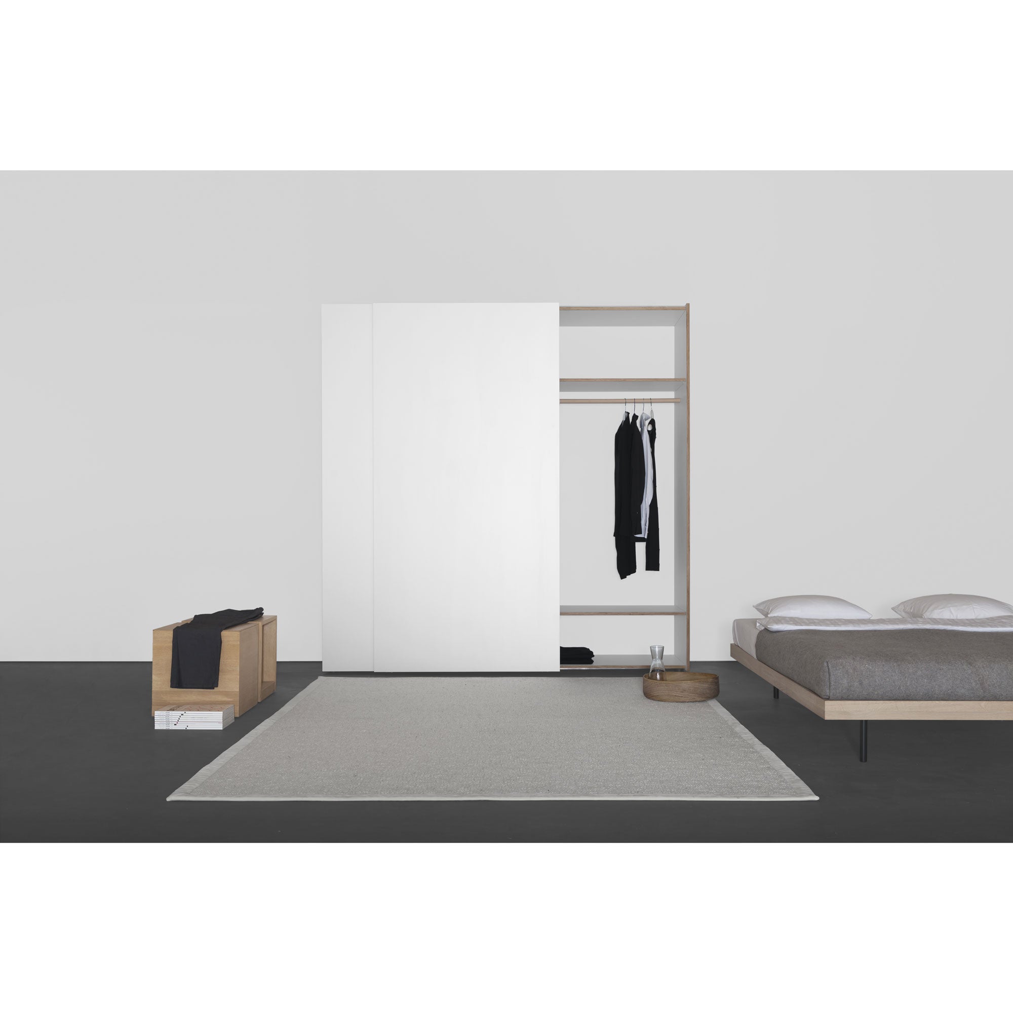 A minimalist bedroom showcases the large Sanktjohanser Kubatur wardrobe with sliding doors, slightly open to display hanging clothes. A low wooden bed with gray bedding is on the right. A centered light gray rug and a small wooden bench with a black item span the left side.