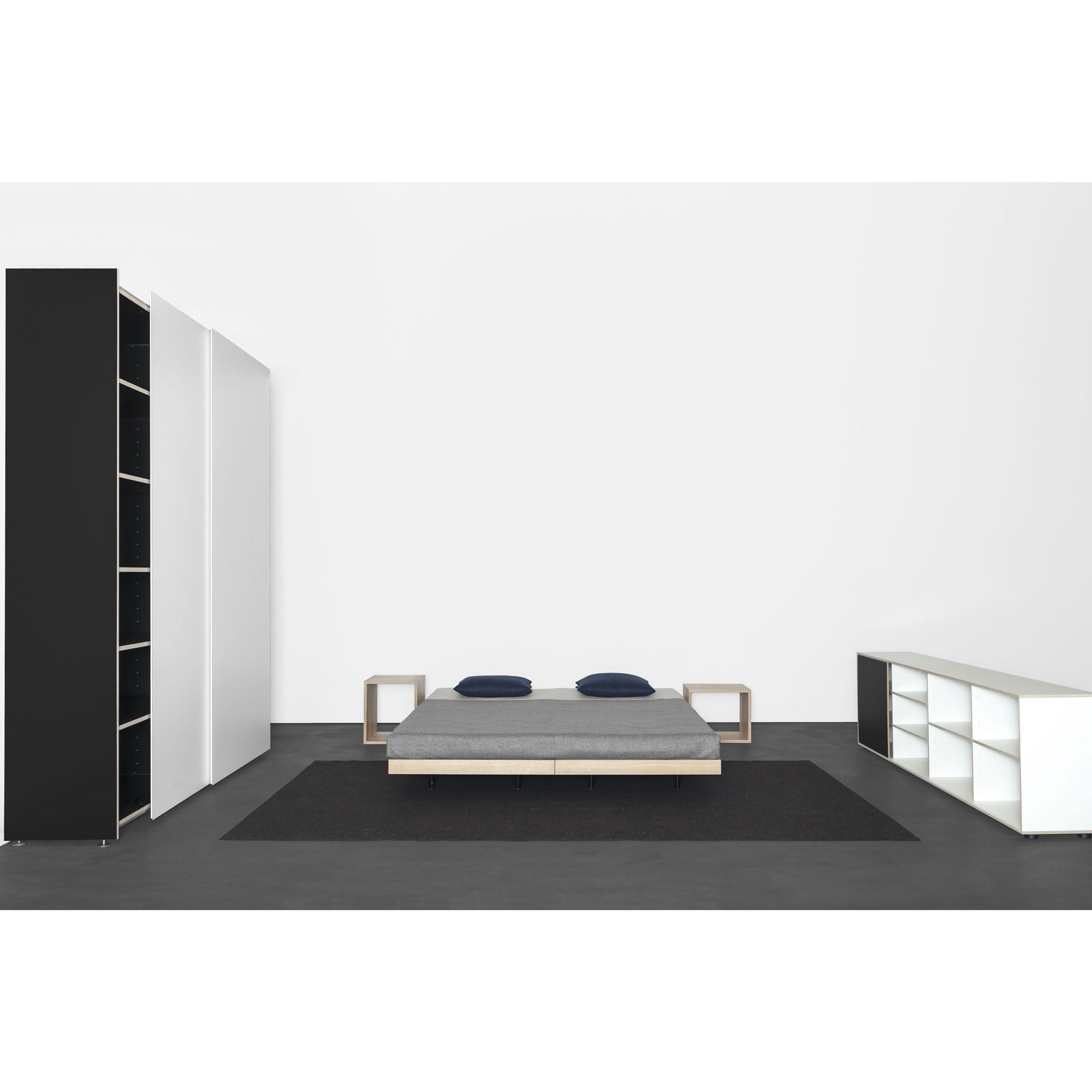 A minimalist bedroom features a low grey bed, two dark pillows, matching side tables on a black rug, and Sanktjohansers Kubatur wardrobe with sliding doors on the left. Its complemented by a white shelving unit on the right, all set on a dark floor against a white wall.