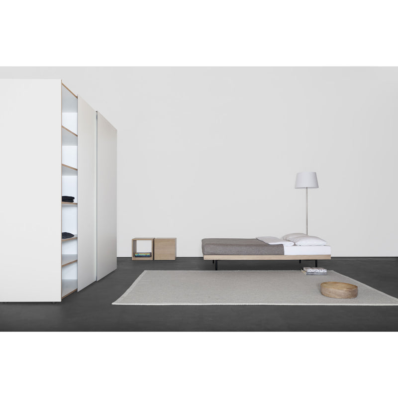 A minimalist bedroom showcases a bed with grey bedding, a wooden cube shelf, and a sleek white floor lamp. Discover the Kubatur wardrobe by Sanktjohanser with open customizable shelves and folded clothes. A small round cushion sits on a light grey rug against the dark solid wood floor.