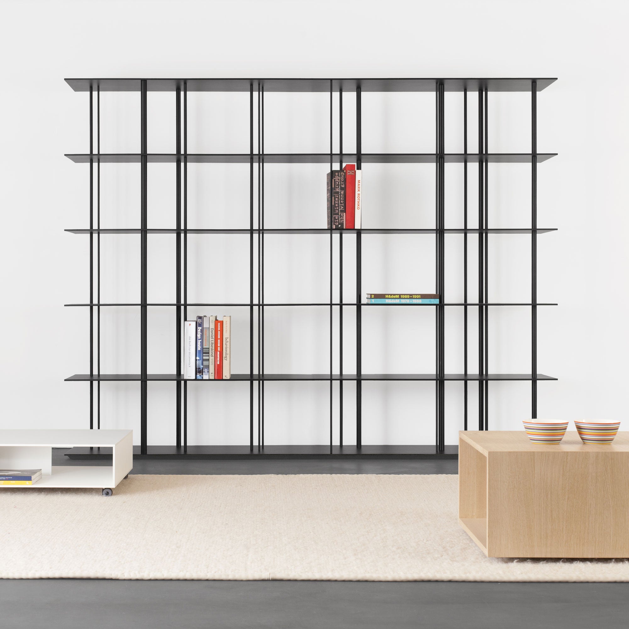 A minimalist room features a black Sanktjohanser Linux shelf against a white wall, holding a few books. A white coffee table and wooden table with two bowls rest on a beige rug, while the dark gray floor contrasts starkly with the light decor.