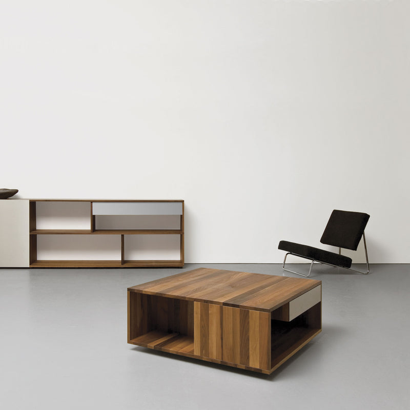 In a minimalist room, the Sanktjohanser Loft coffee table, modern low chair with black cushions, and a wooden sideboard with open shelves against a white wall create an open, uncluttered, and calm atmosphere.