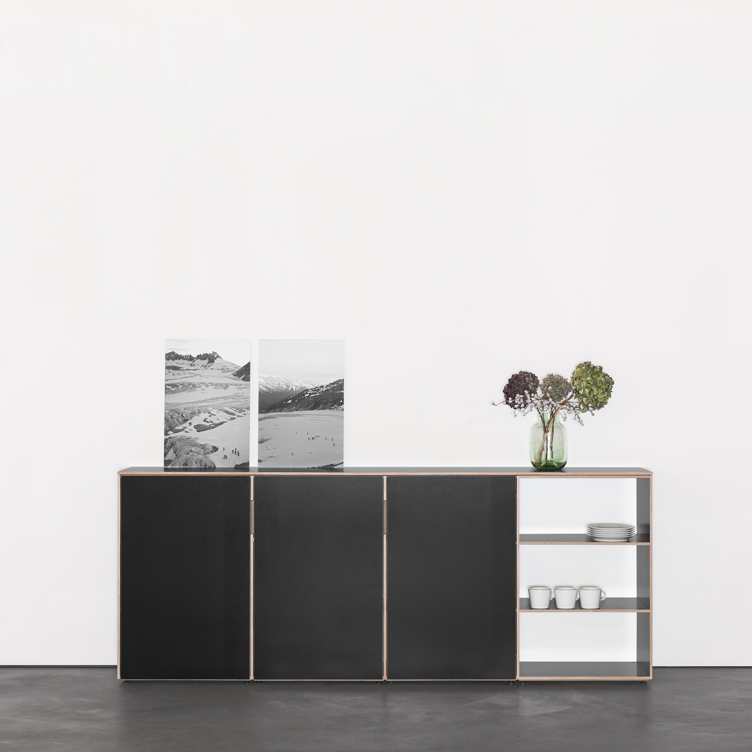 The Metrum Sideboard by Sanktjohanser is a minimalist black, made-to-measure cabinet against a white wall with two black-and-white landscape photos. A vase of dried flowers and three stacked white bowls on open shelves add decoration.