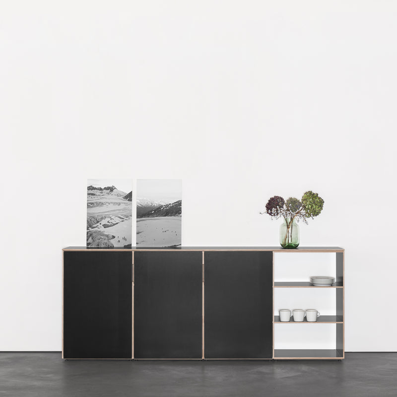 The Metrum Sideboard by Sanktjohanser is a minimalist black, made-to-measure cabinet against a white wall with two black-and-white landscape photos. A vase of dried flowers and three stacked white bowls on open shelves add decoration.