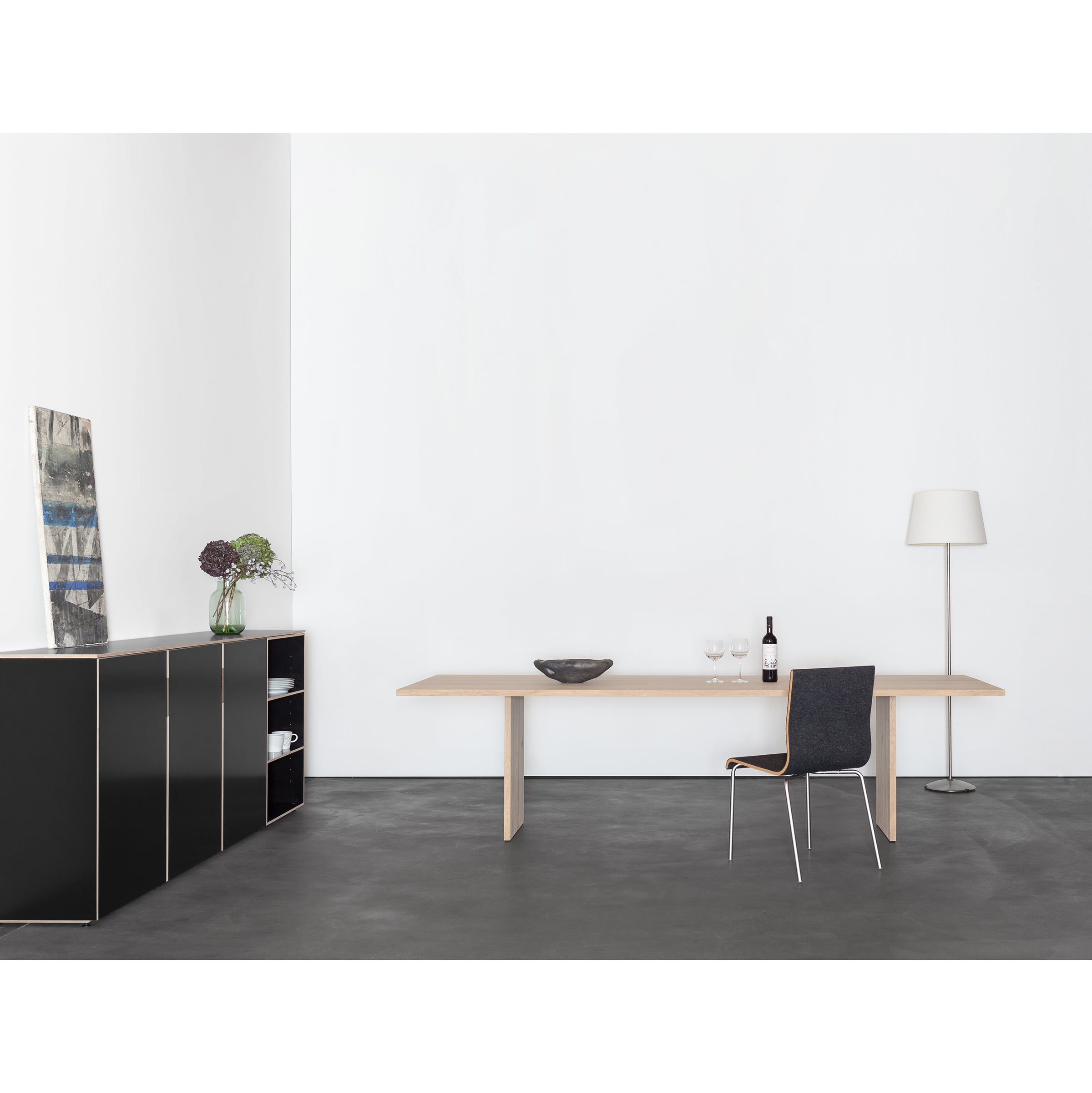 Experience the minimalist elegance of a room with a Sanktjohanser Alphabet Table, showcasing solid wood finishes. Complemented by a chair, wine bottle, standing lamp, and black cabinet adorned with plants and art, this setup features the unique charm of 26 distinctive leg designs.