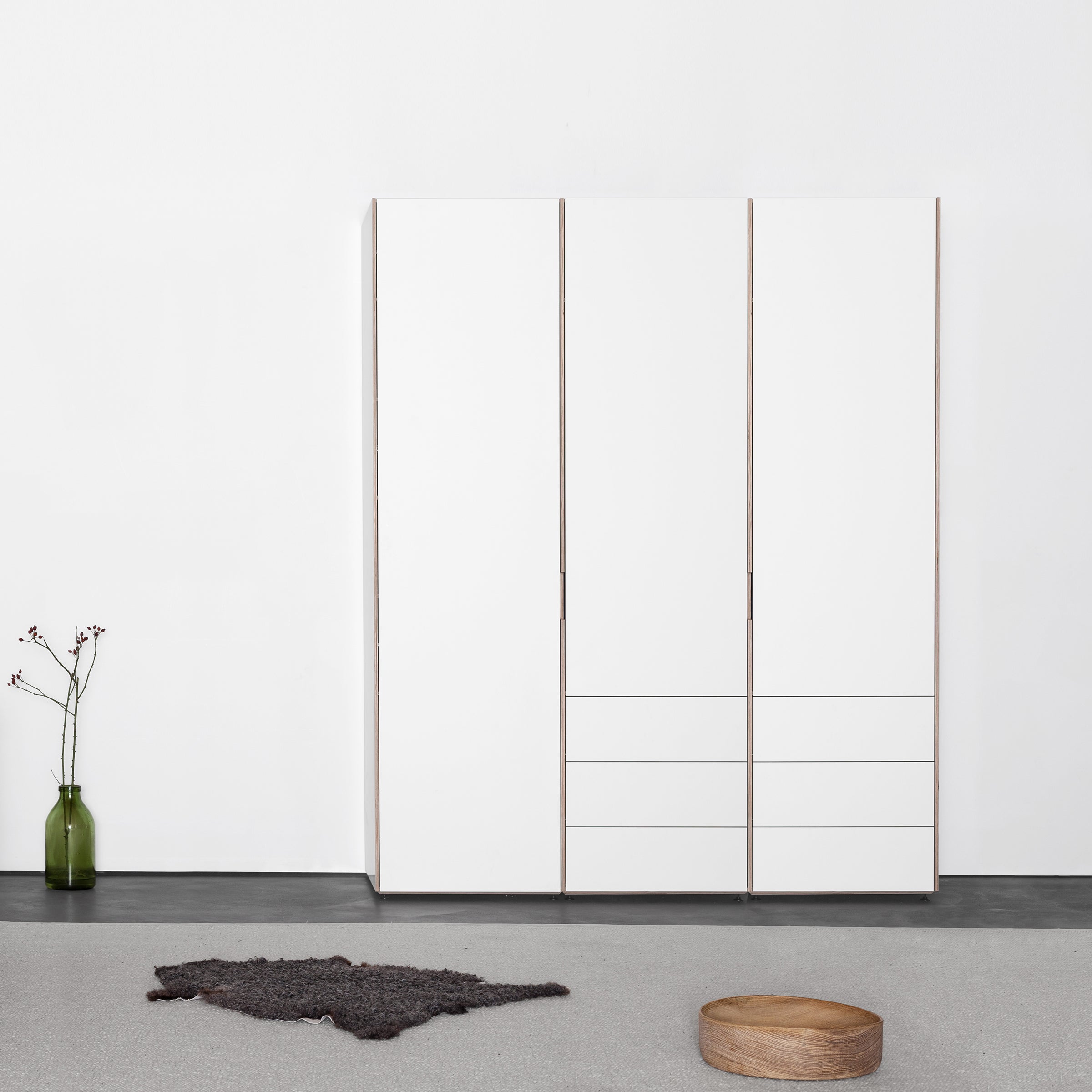 A minimalist room features a large white Sanktjohanser Metrum Storage cabinet seamlessly integrated into the wall. To the left, a small green vase with flowers contrasts with the black rug below and complements the nearby round wooden stool, creating a harmonious space.