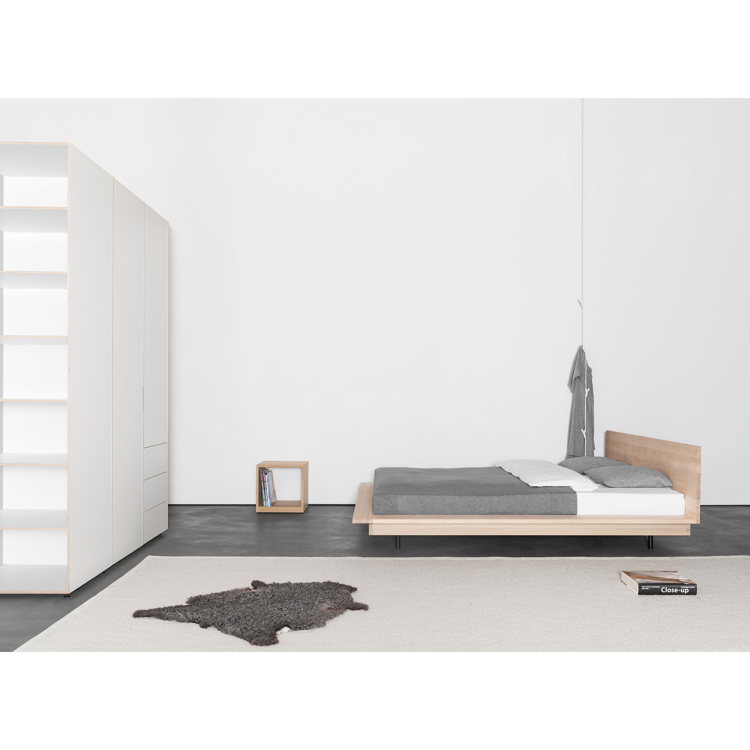 A minimalist bedroom with a low wooden bed and gray bedding features the Sanktjohanser Metrum Storage cabinet on the left for an elegant storage system. A small wooden bedside table holds books, while a light rug is on the floor and a robe hangs on the wall.