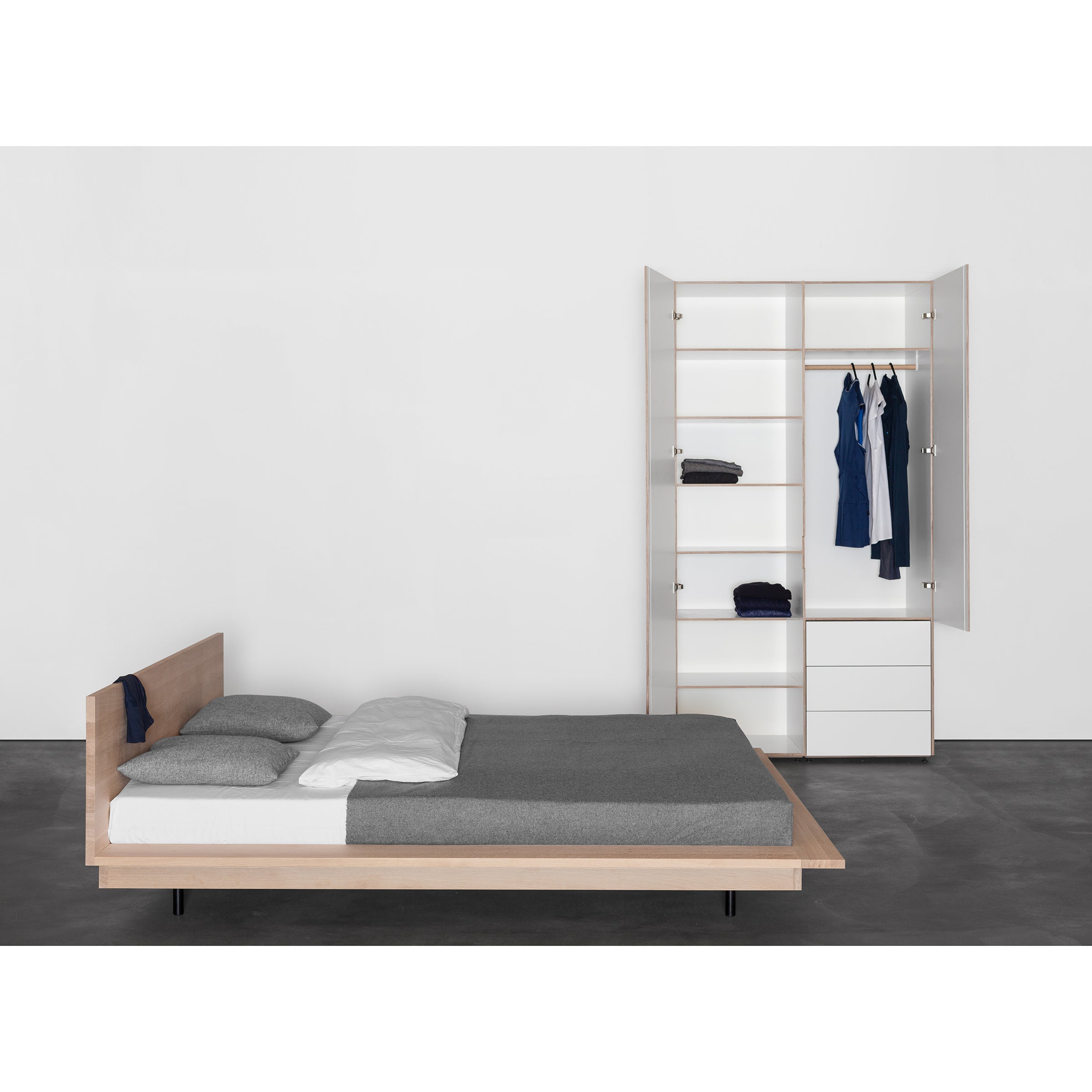 A minimalist bedroom showcases a wooden bed frame, gray bedding, and the Metrum Storage wardrobe by Sanktjohanser with open doors. Neatly arranged clothes are set against a plain white wall on a dark gray floor, exemplifying made-to-measure elegance.