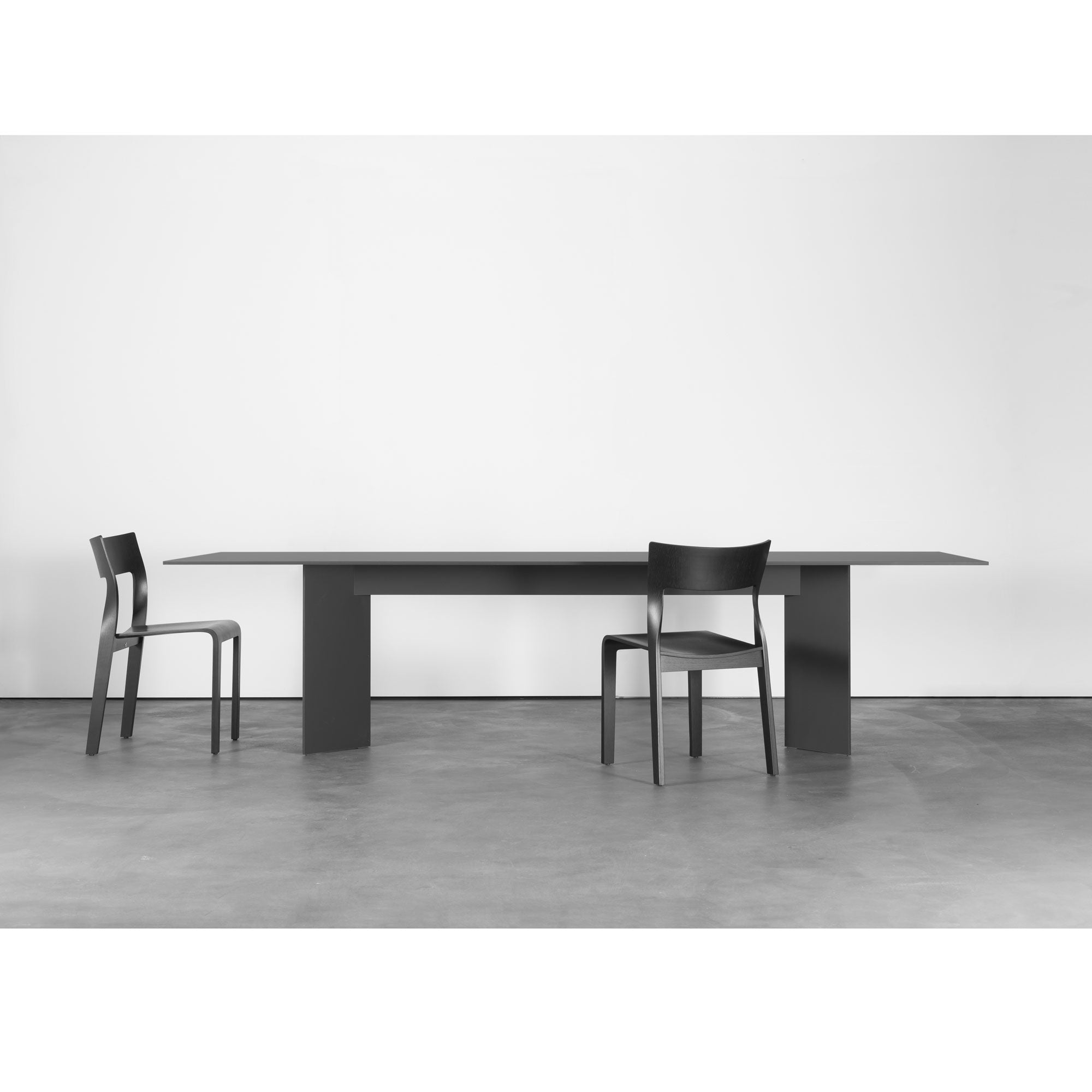 A minimalist room features a sleek Sanktjohanser Mono Table, complemented by two black chairs at opposite ends. The stark white wall and smooth concrete floor highlight the tables clean, modern design, making it a stylish and customizable centerpiece.