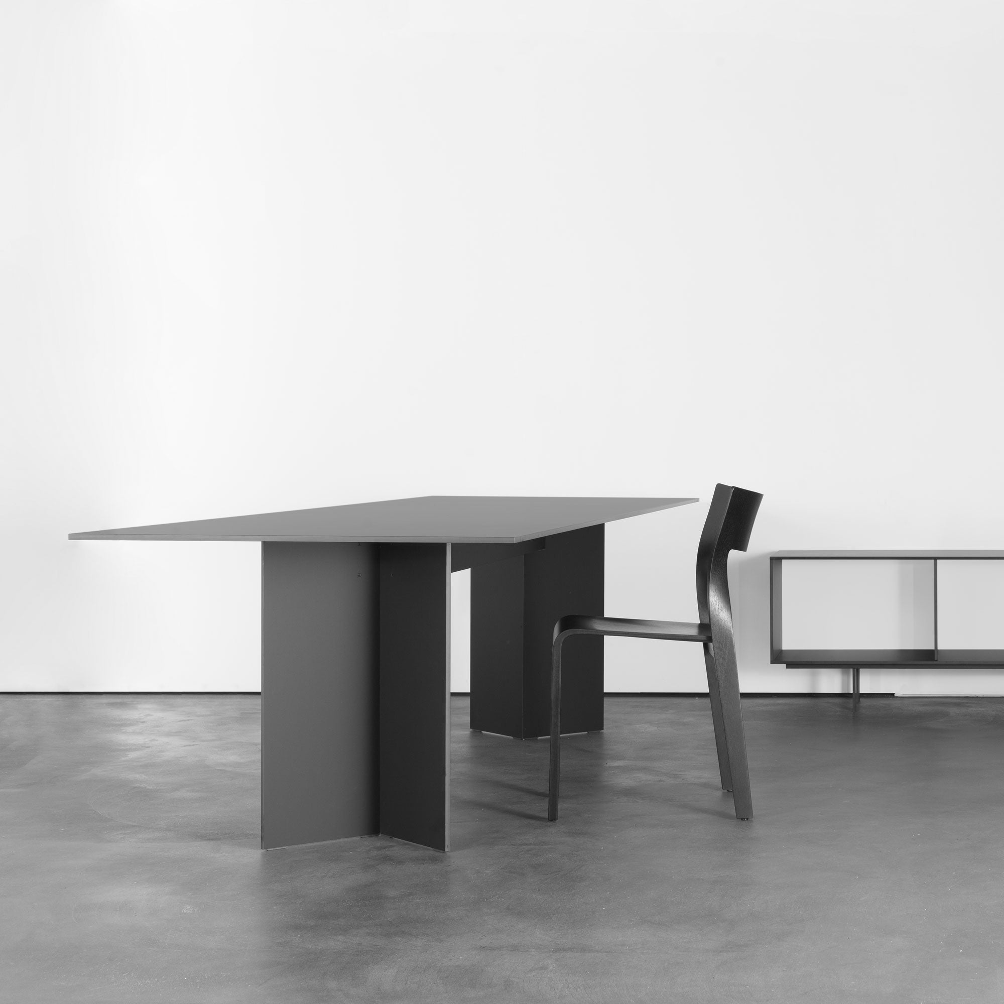 Minimalist room with a sleek Sanktjohanser Mono Table in black, featuring a geometric base and paired with a matching chair and low cabinet against a plain white wall, all set on a smooth gray concrete floor.
