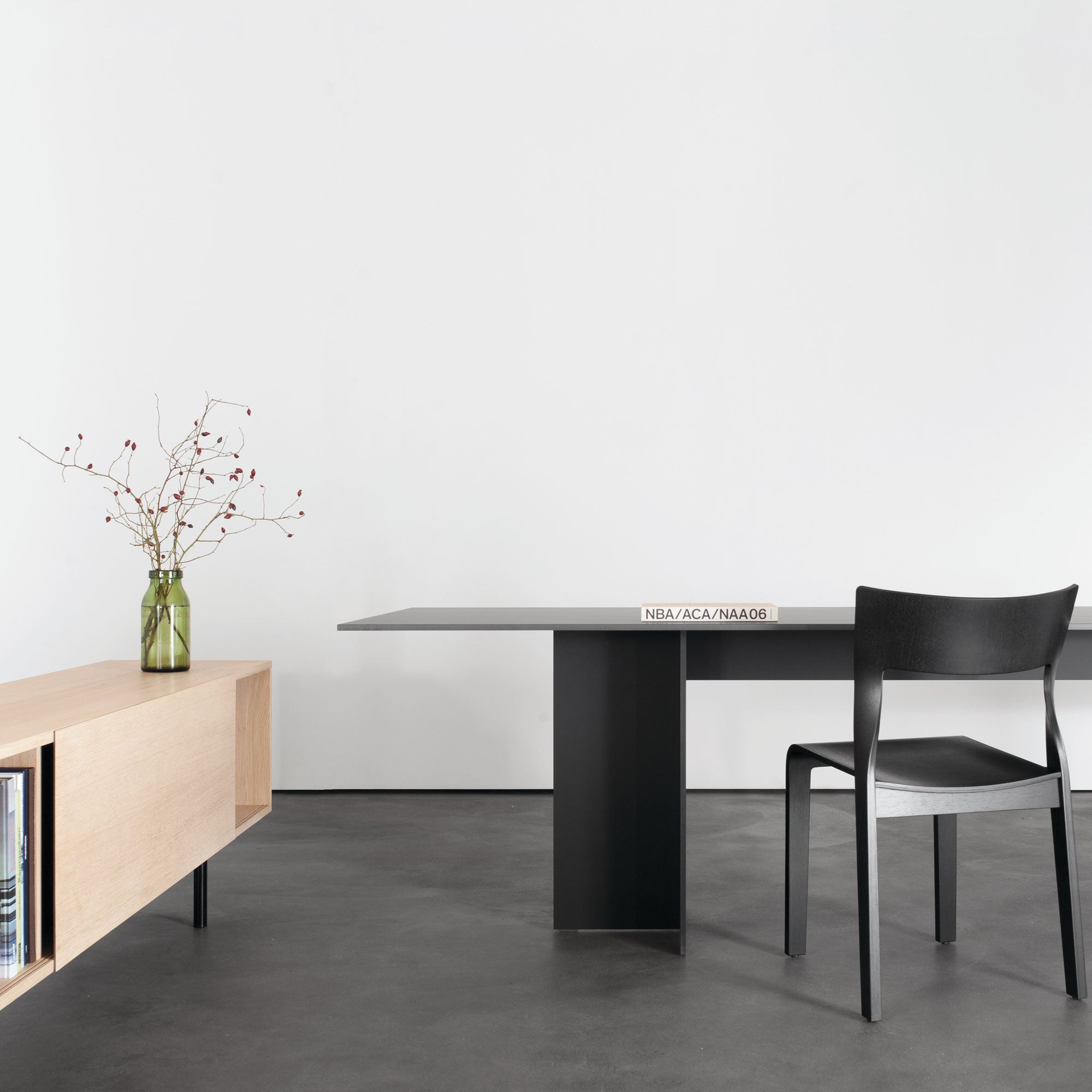 A minimalist room with a black chair and a Sanktjohanser Mono Table on dark flooring. A light wood sideboard supports a green vase with branches. The contemporary design, featuring neutral tones, is enhanced by the customizable tables versatility in creating an elegant space.