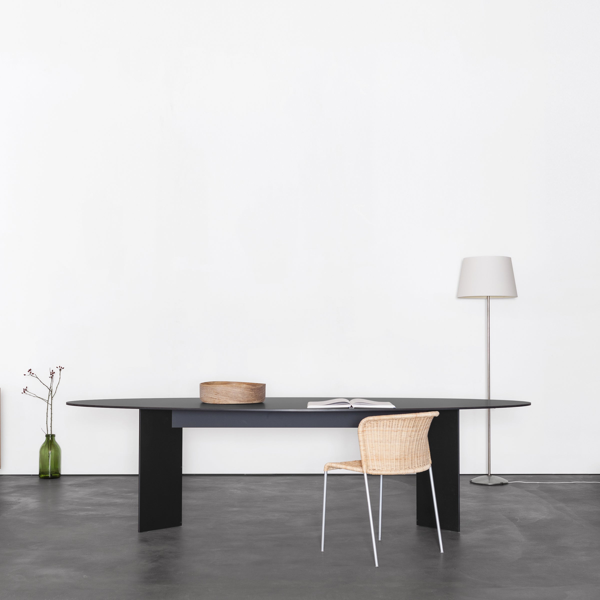 A minimalist room showcases a large black Mono Table Oval by Sanktjohanser, paired with a wicker chair and an open book. Nearby, a tall floor lamp highlights a green vase with branches on the white wall. The concrete floor completes this Fenix-inspired space.
