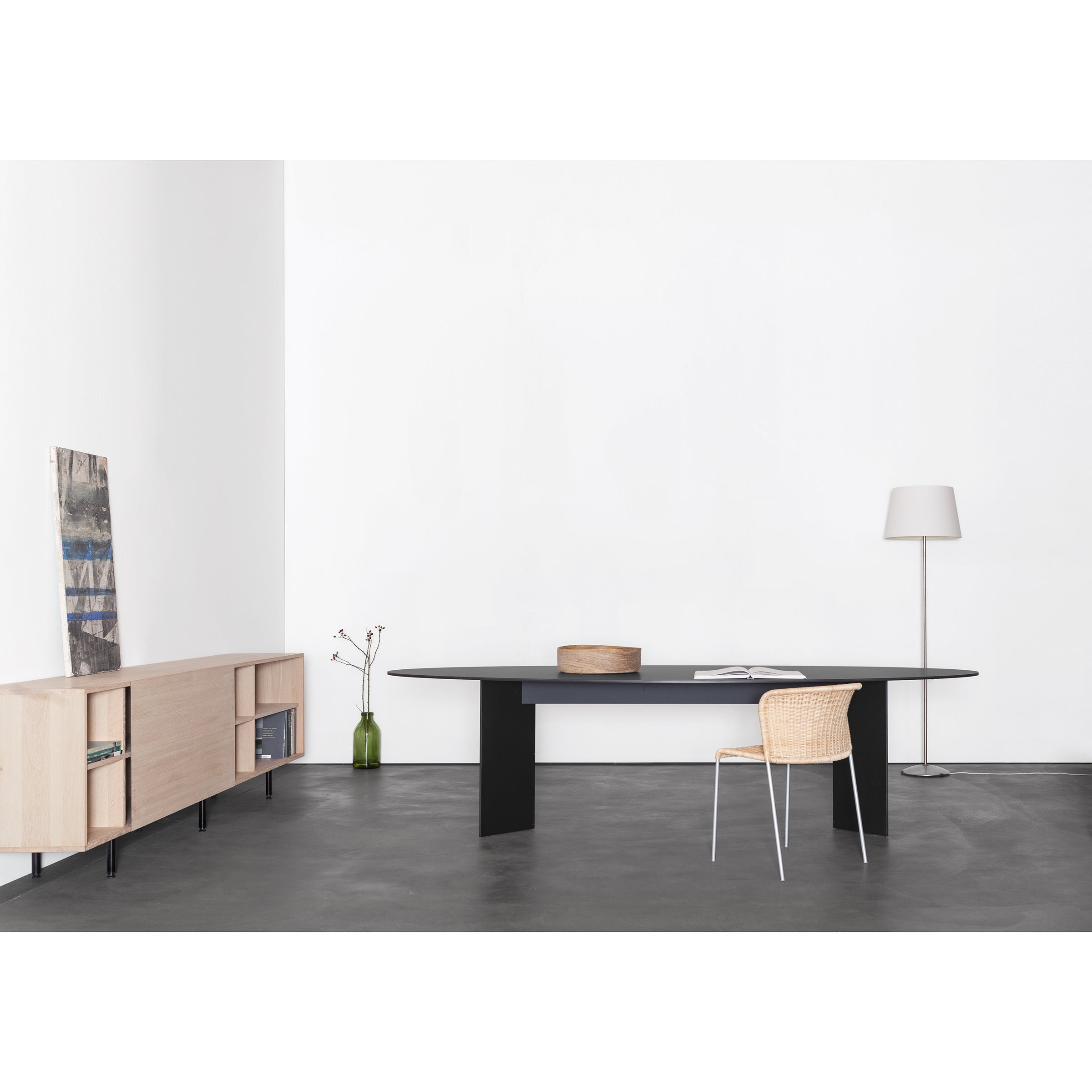 In a minimalist room, the sleek black Mono Table Oval by Sanktjohanser complements a wicker chair on the dark floor. A light wood Sanktjohanser sideboard on the left holds books and a vase, while a tall floor lamp stands in the corner against plain white walls.
