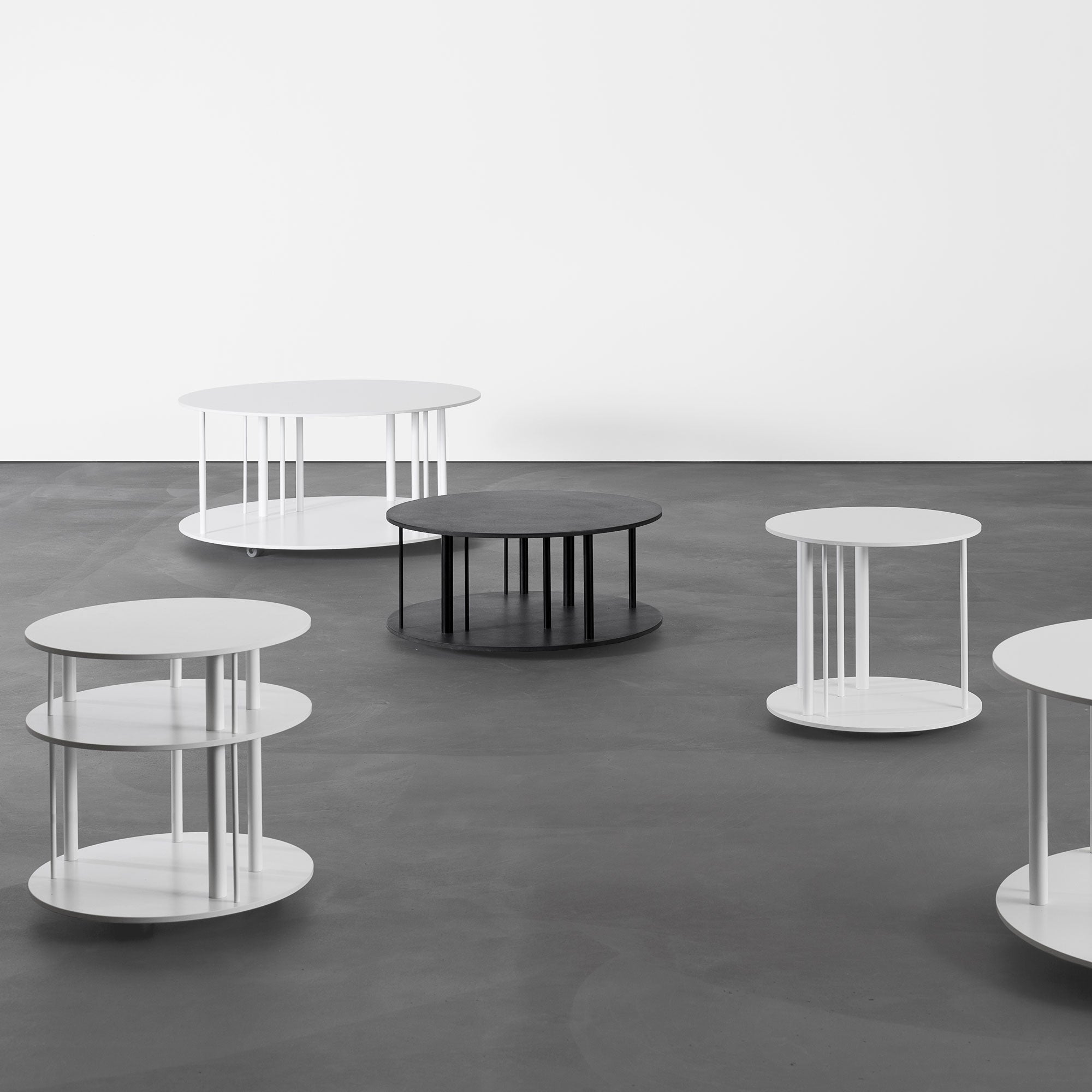 The minimalist interior showcases six round Pi Coffeetables by Sanktjohanser: five white and one black, crafted from solid wood. Each unique design features two circular surfaces linked by vertical rods, all set against a smooth gray floor and plain white wall.