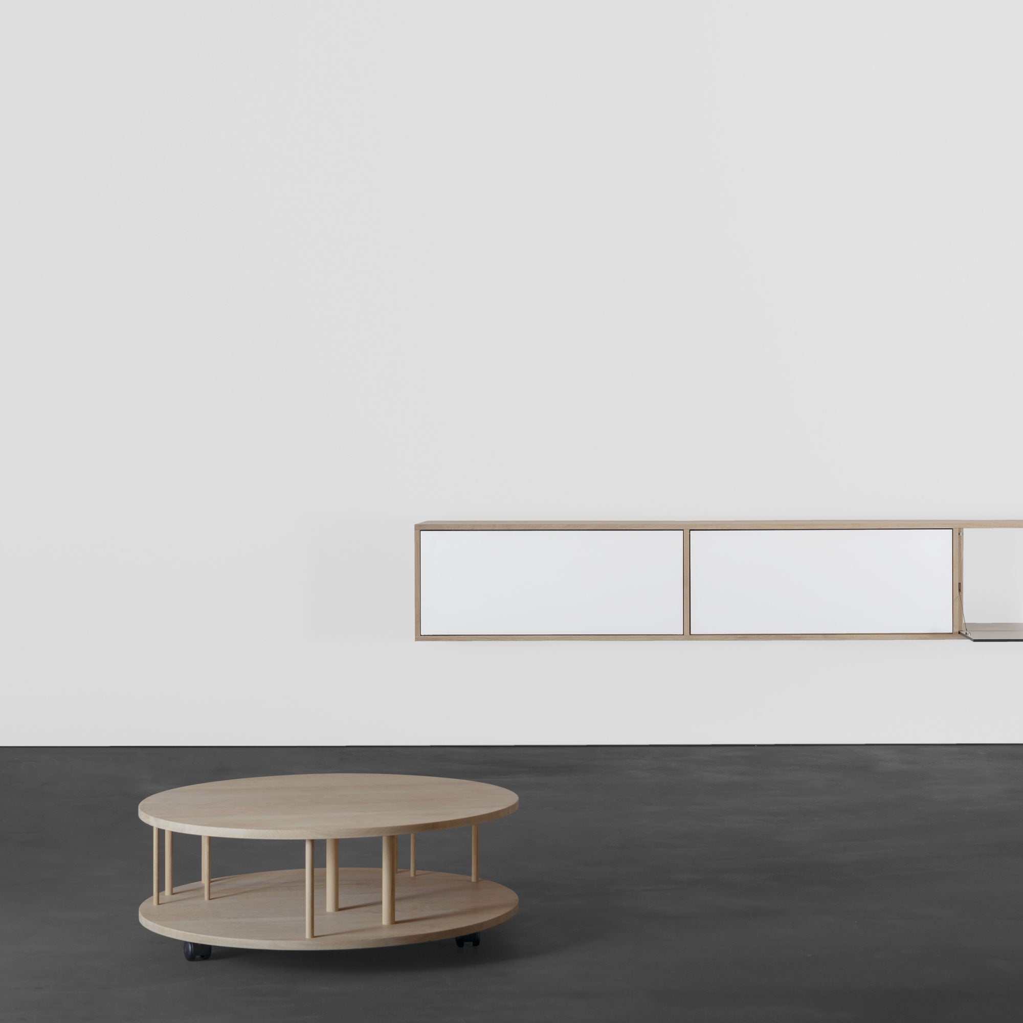 In a minimalist room, the Pi Coffeetable by Sanktjohanser sits on wheels against a dark floor. A white wall in the background features a distinctive design with an elegantly mounted low, rectangular shelving unit.