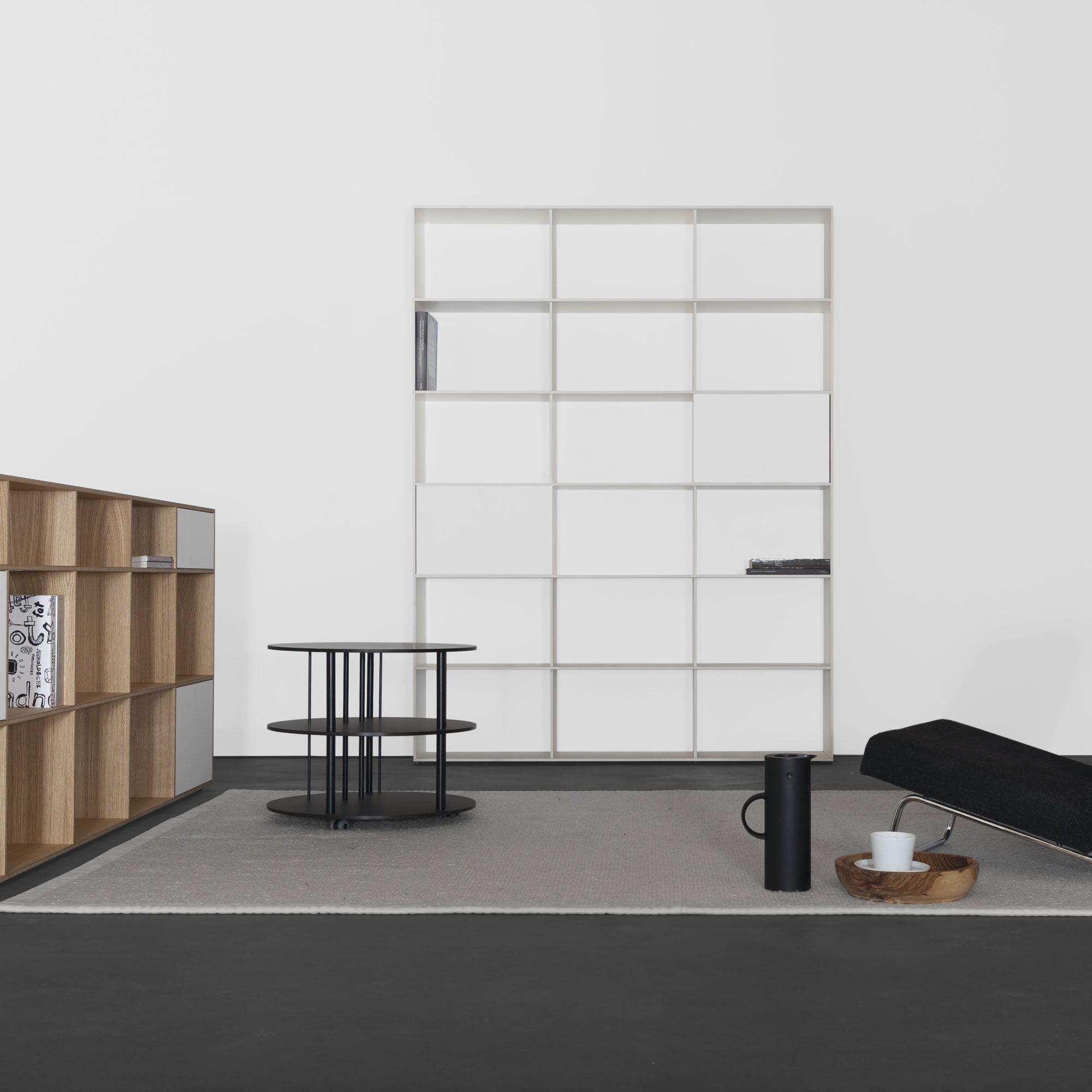 A minimalist room features a black sofa and the uniquely designed Pi Coffeetable by Sanktjohanser. Wooden and white storage shelves complement the grey rug on a dark floor. A black carafe and cup sit on a wooden tray, adding hints of solid wood elegance to the rooms charm.
