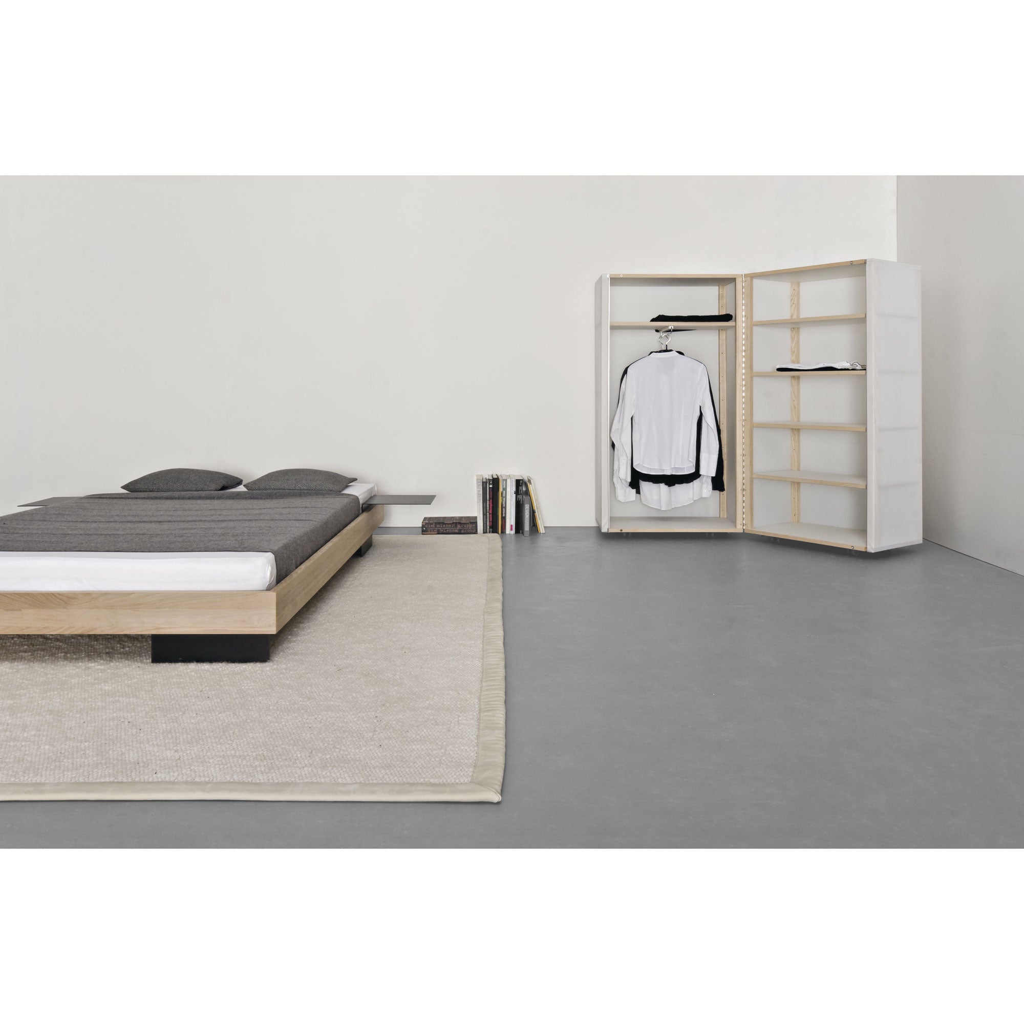 A minimalist bedroom features a low wooden bed, gray bedding, and a beige rug. The room has an open Quartier wardrobe by Sanktjohanser with translucent panels showcasing a hanging white shirt and jacket, flanked by shelves of books on the right, offering a simple, modern aesthetic.