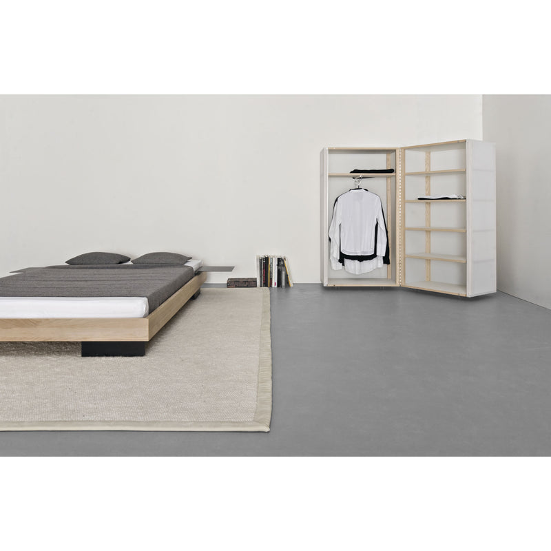 A minimalist bedroom features a low wooden bed, gray bedding, and a beige rug. The room has an open Quartier wardrobe by Sanktjohanser with translucent panels showcasing a hanging white shirt and jacket, flanked by shelves of books on the right, offering a simple, modern aesthetic.