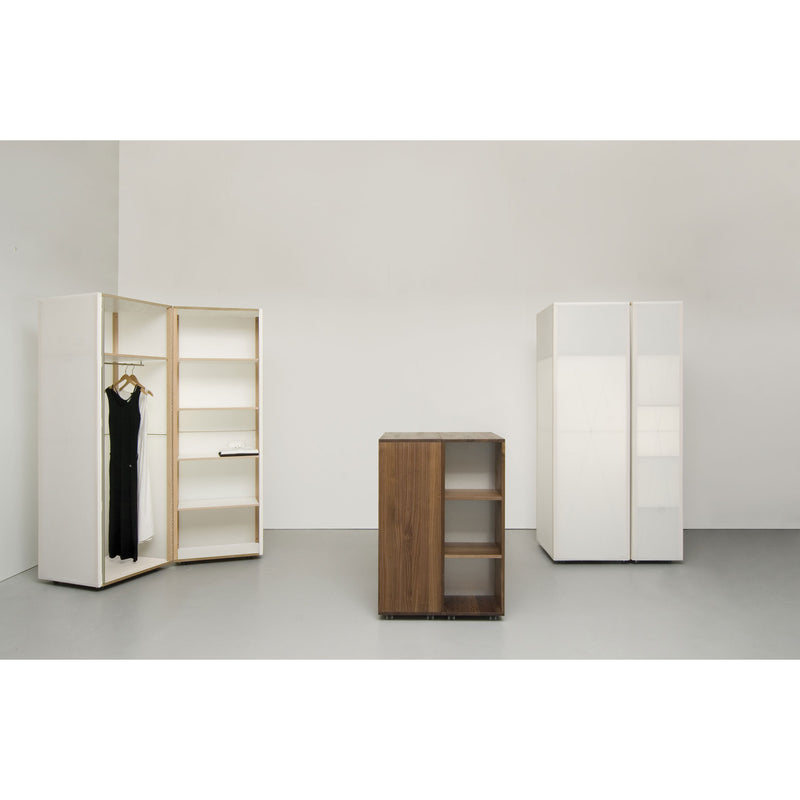 A minimalist room showcases a translucent Quartier wardrobe by Sanktjohanser next to an open white wardrobe with shelves holding a black dress. A small solid oak shelving unit centers the space, while the walls and floor maintain a neutral gray tone.