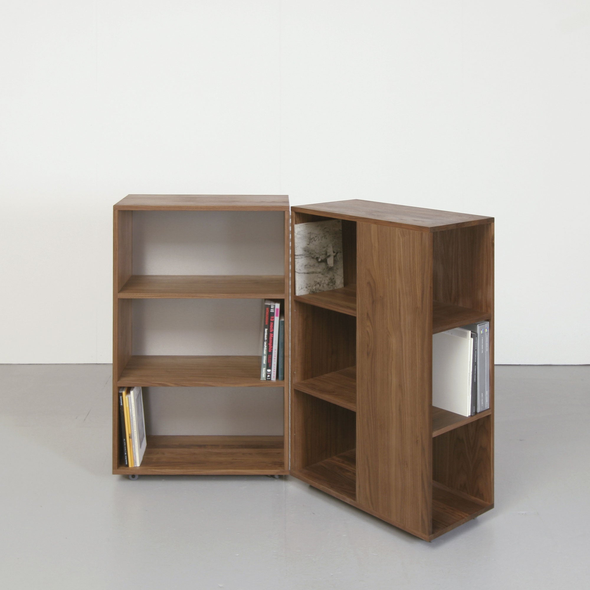 Two Quartier wardrobes by Sanktjohanser, with solid oak frames, stand on a light gray floor against a white wall. Each has three open shelves, sparsely filled with books on the bottom and middle levels. The left wardrobe is slightly angled towards the viewer.
