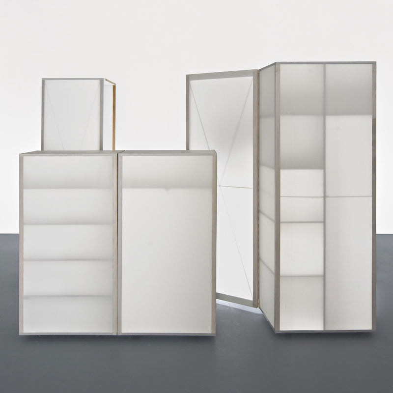 The Quartier wardrobe by Sanktjohanser features minimalist white cabinets with a solid oak frame, resting elegantly on a dark gray floor. Some doors ajar reveal geometric designs through transparent and opaque panels, creating a sleek look against the plain white backdrop.