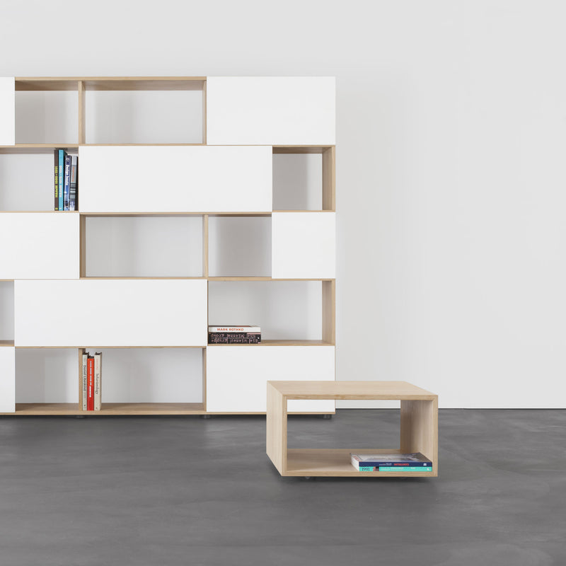 A minimalist room features modular furniture with the Relikt multipurpose bookshelf by Sanktjohanser, showcasing asymmetrical white panels. A solid wood side table holds books, set against a gray floor and plain white wall.