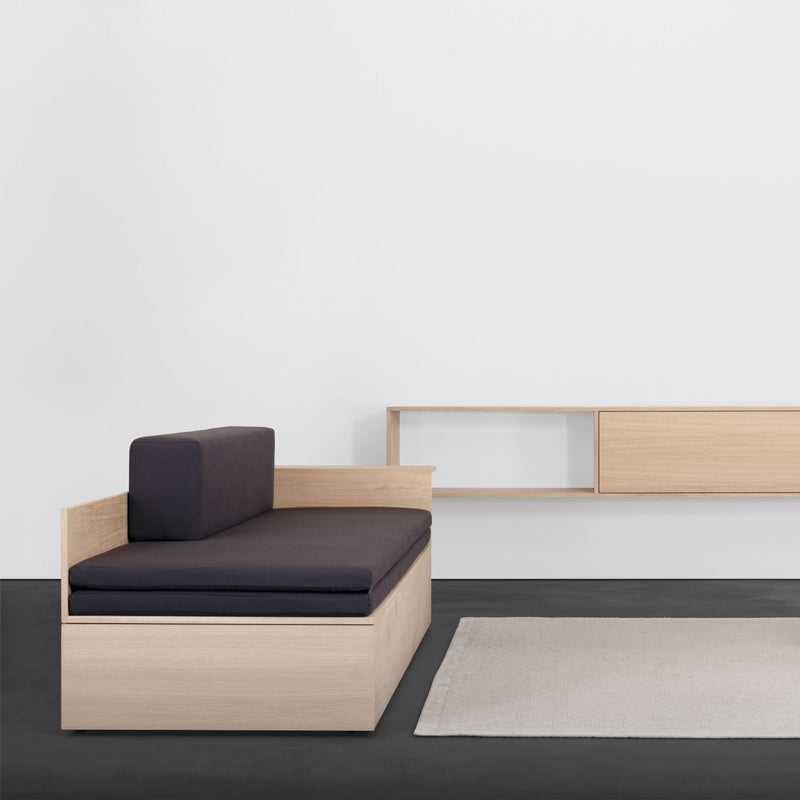 A minimalist room features a Sanktjohanser Salto Daybed with black cushions, paired with a light wood floating shelf on the white wall and a beige rug on the dark floor, emphasizing clean lines and simplicity in its design.