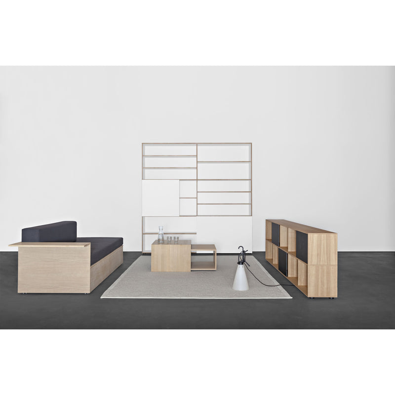 In a minimalist living room, a low solid wood sofa and matching coffee table rest on a gray rug. Light wood furniture, including a shelving unit, complements the sleek Sanktjohanser Salto Daybed next to the floor lamp. A glass of water sits on the table amid geometric wall art in the background.