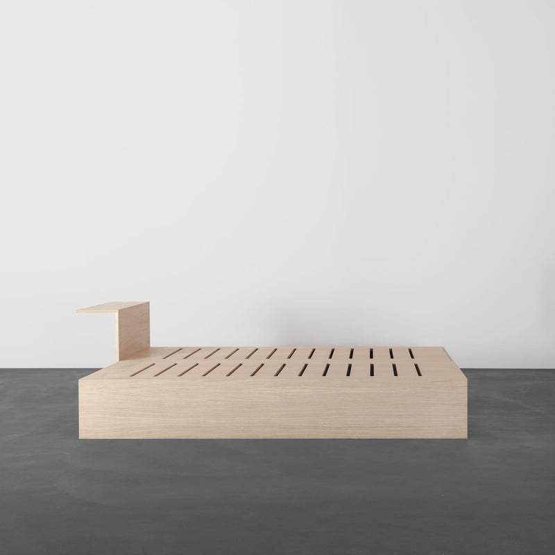 The Sanktjohanser Salto Daybed boasts a minimalist design with a light solid wood finish, featuring a unique slotted frame and attached elevated side table, set against a plain white wall on a dark floor.