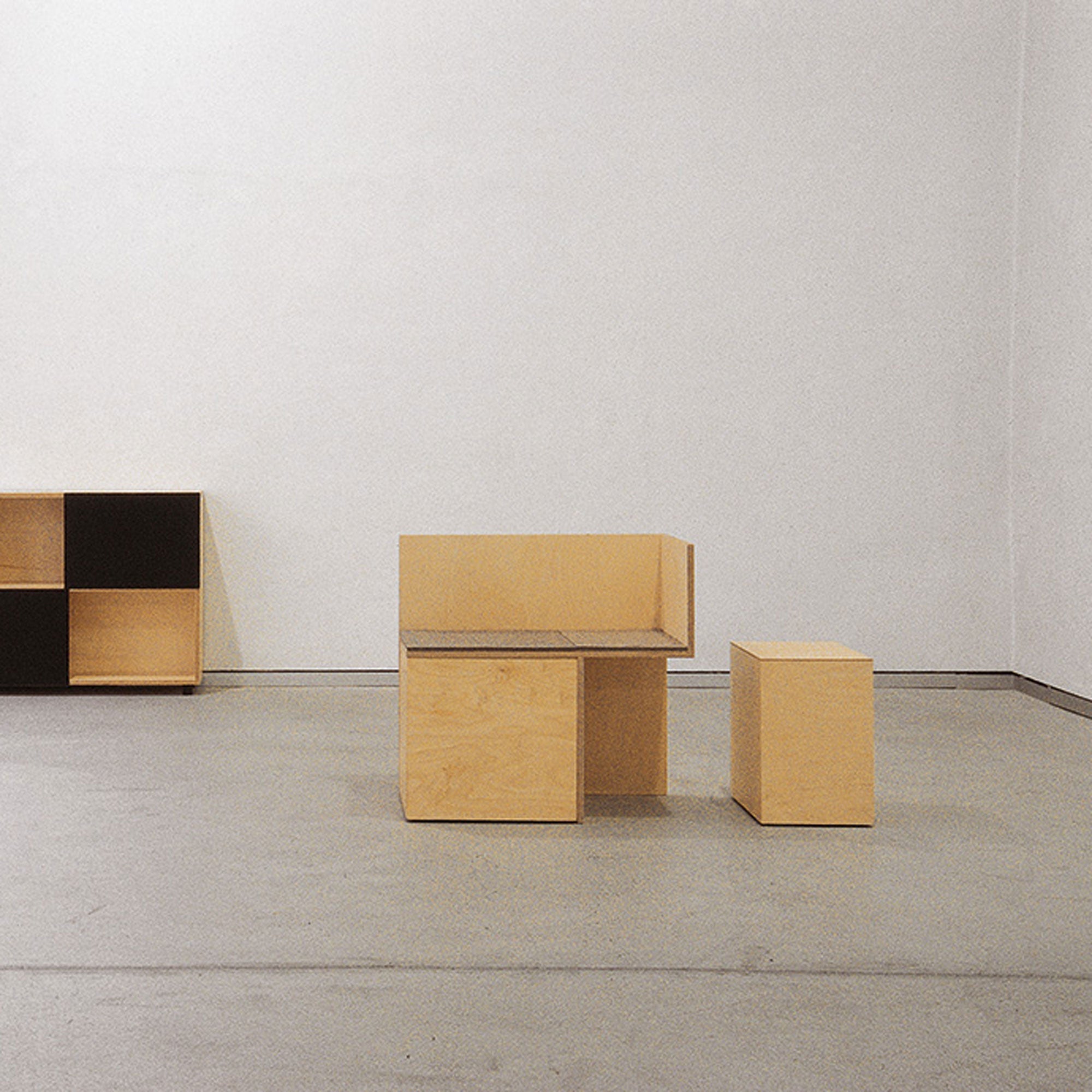 A minimalist room features light wood furniture against a white wall, highlighted by a geometric chair and table set. A black and wood shelving unit on the left showcases the versatile Social Cube system by Sanktjohanser. The concrete floor complements the sparse, modern aesthetic.