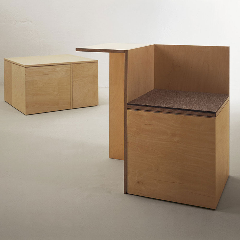 The Sanktjohanser Social Cube includes a minimalist wooden chair with a backrest and side table, a dark cushioned seat, and an adaptable wooden storage cube. The set sits on a light-colored floor.