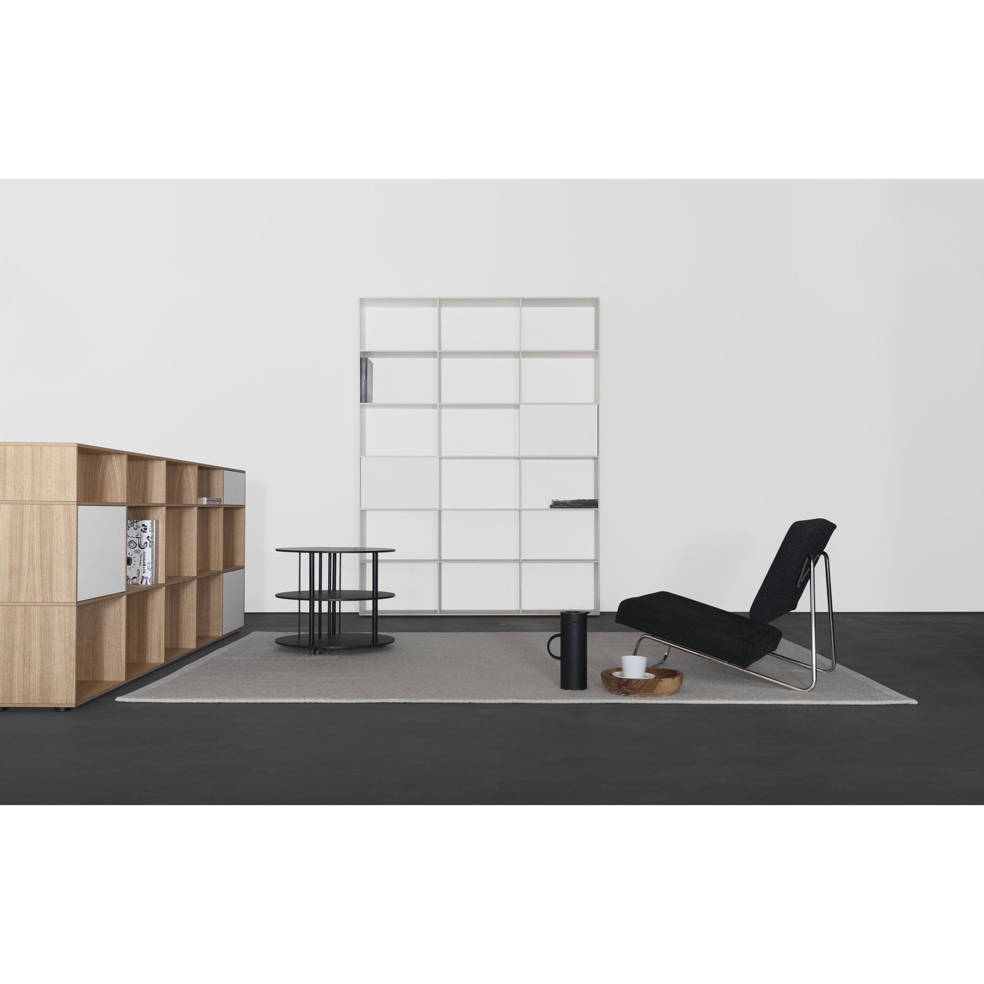 A modern minimalist room features a black lounge chair, a round coffee table, a gray rug, the customizable Tektur sideboard from Sanktjohanser in oak veneer, and grid-patterned artwork against a white wall. The dark floor contrasts with the light decor elements.