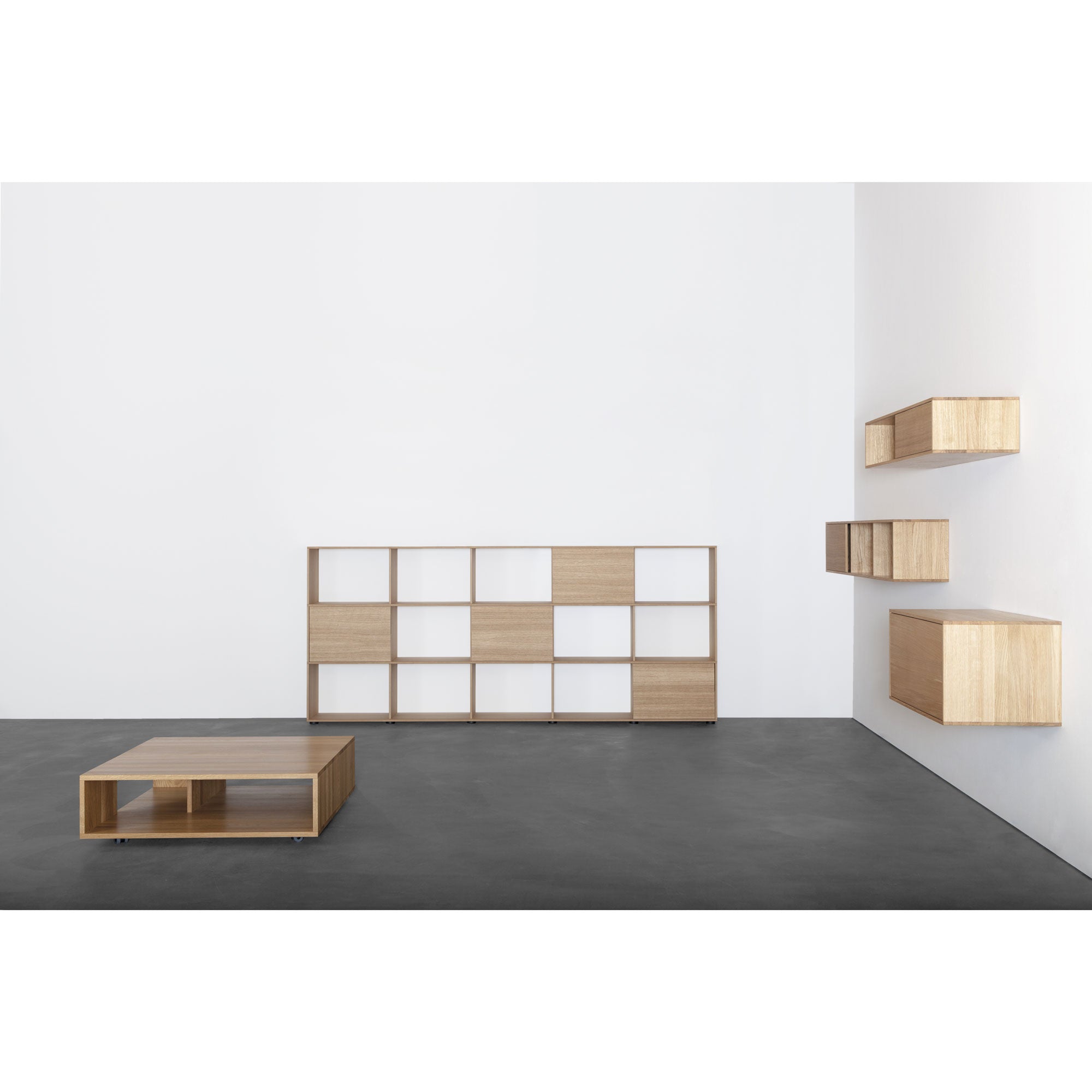 A minimalist room features light wood furniture, oak veneer accents, white walls, and dark flooring. It includes a low coffee table, a large customizable shelf system with mixed open/closed sections, three floating wall cabinets, and the Tektur sideboard by Sanktjohanser.