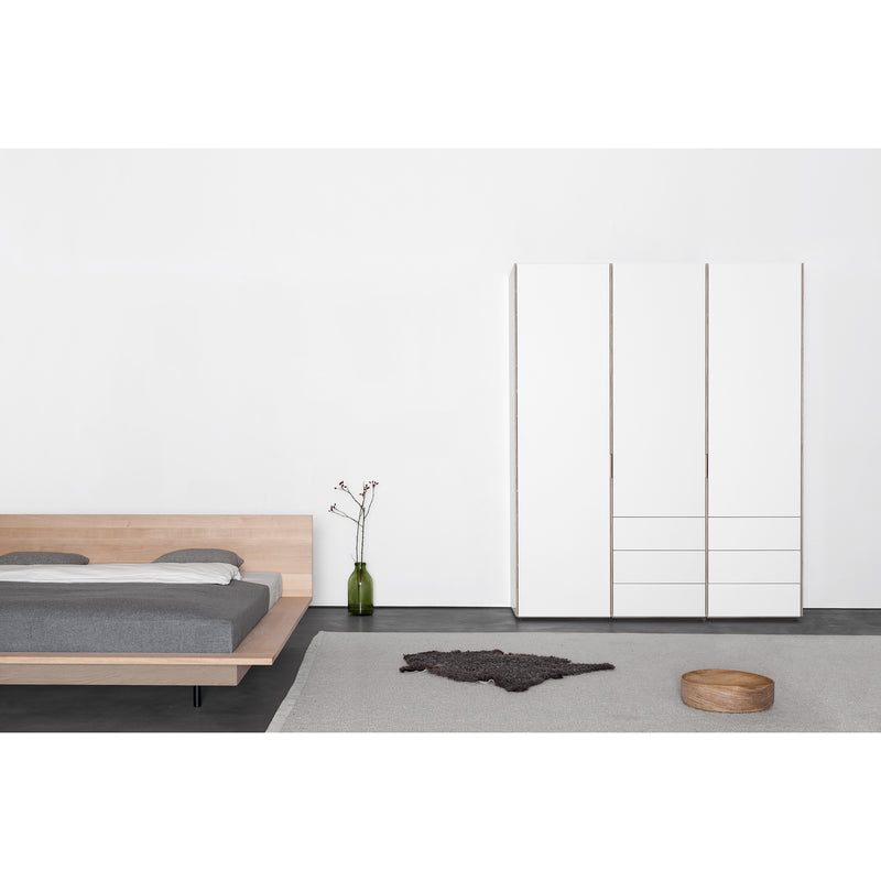 The minimalist bedroom showcases a customizable Unisono Bed by Sanktjohanser with a solid wood frame and gray bedding. A white wardrobe is against the wall. A green vase decorates the bedside, and a circular wooden tray is on a light gray rug next to the dark one.