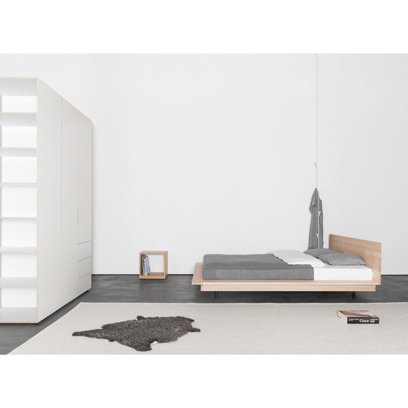 The minimalist bedroom showcases a low platform Sanktjohanser Unisono bed with gray bedding. A gray robe is on the right, a white wardrobe on the left, and beside the solid wood bed, a small shelf holds books. A textured gray rug on light carpet ties the room together seamlessly.