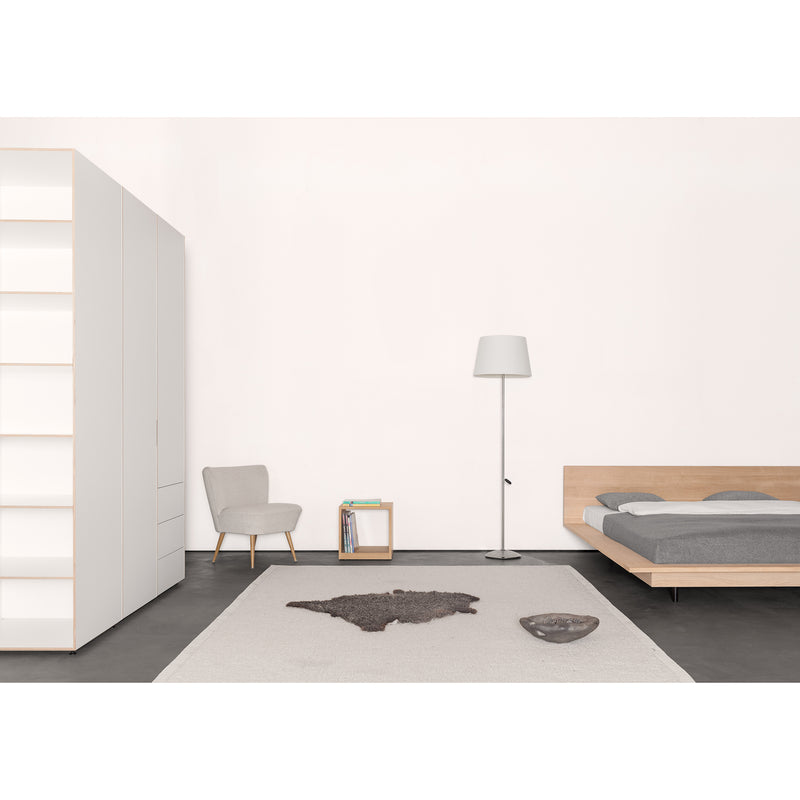 A minimalist bedroom showcases a Sanktjohanser Unisono bed with gray bedding, a white lamp, and a customizable wardrobe. A square shelf holds a book beside the small chair. A gray bowl and dark mat rest on the light rug against a solid wood-accented white wall.