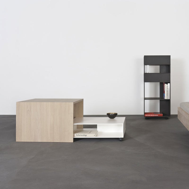 A minimalist room features a Sanktjohanser Matrix multipurpose coffee table with customizable dimensions and a lower shelf for books and bowls, alongside a slim black shelving unit made from HPL material against a white wall on a dark floor.