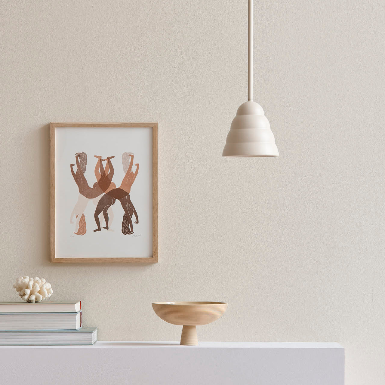 A minimalist interior showcases a framed abstract art piece on a white wall. On the shelf below, stacked books accompany a delicate ceramic bowl. Suspended above is Schneids Dais, a cream pendant light that embodies modern home elegance.