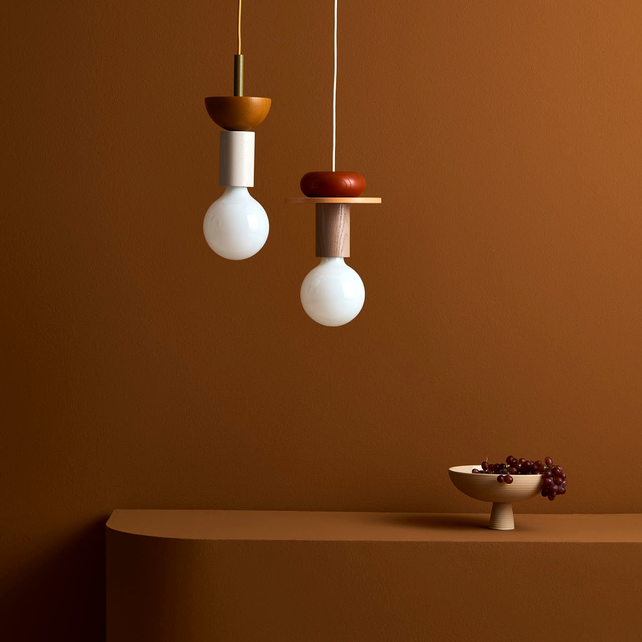 Two Dais pendant lights by Schneid hang against a warm brown wall. On the matching ledge below sits a minimalist ceramic bowl with grapes, creating a harmonious and elegant composition perfect for modern homes.